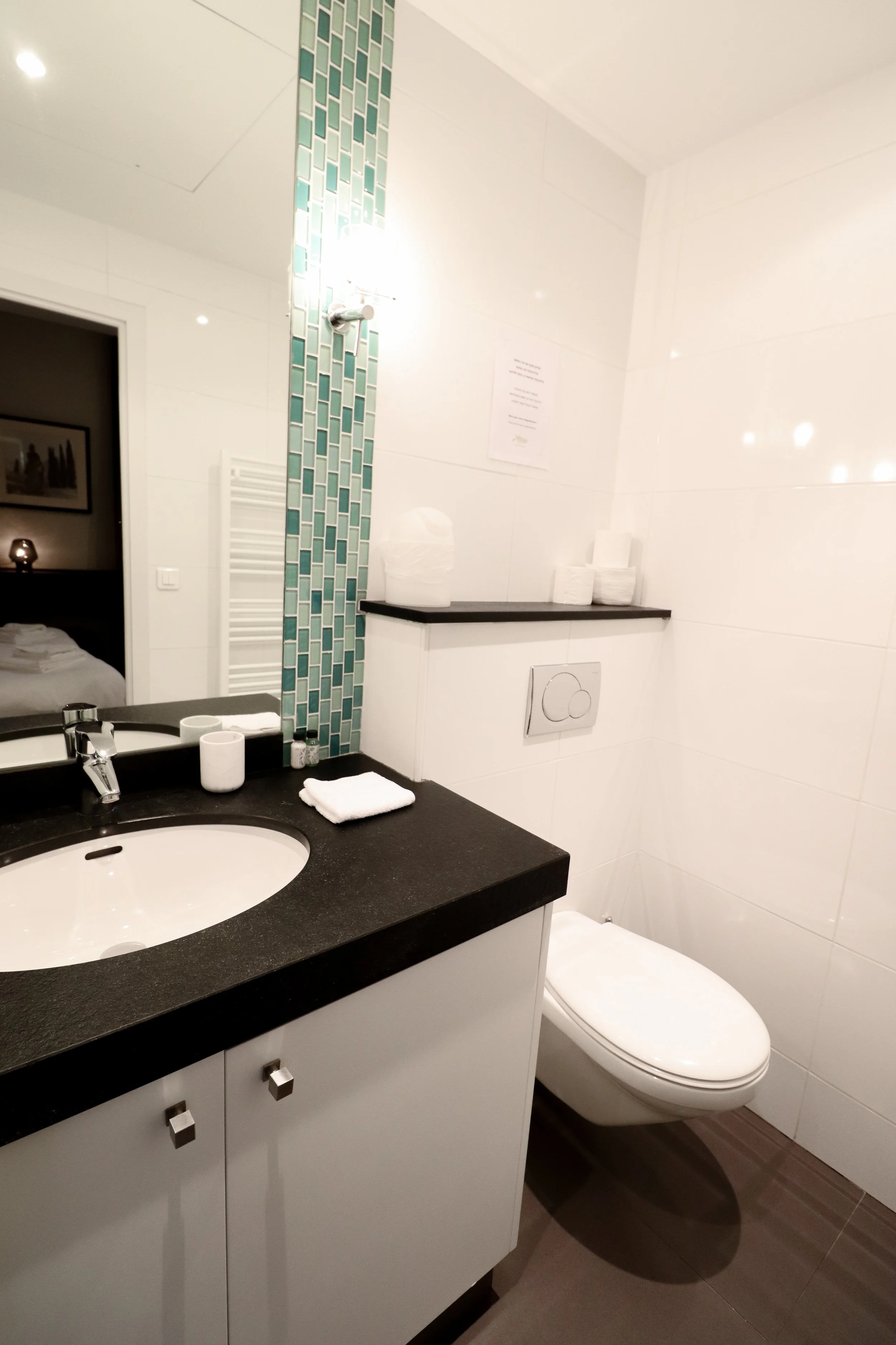 Salle de bains avec un lavabo noir, un miroir, un WC et des serviettes. Mosaïque bleu-vert et murs blancs.
