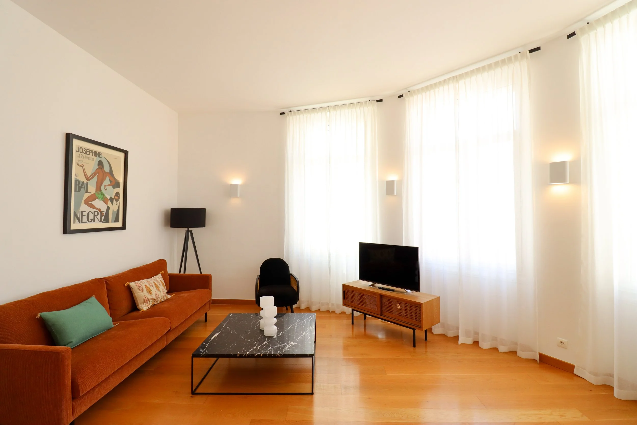 Salon avec un canapé orange, table basse en marbre noir, télévision sur meuble en bois, fenêtres avec rideaux blancs, tableau coloré au mur, lampe noire et petite décoration sur la table.
