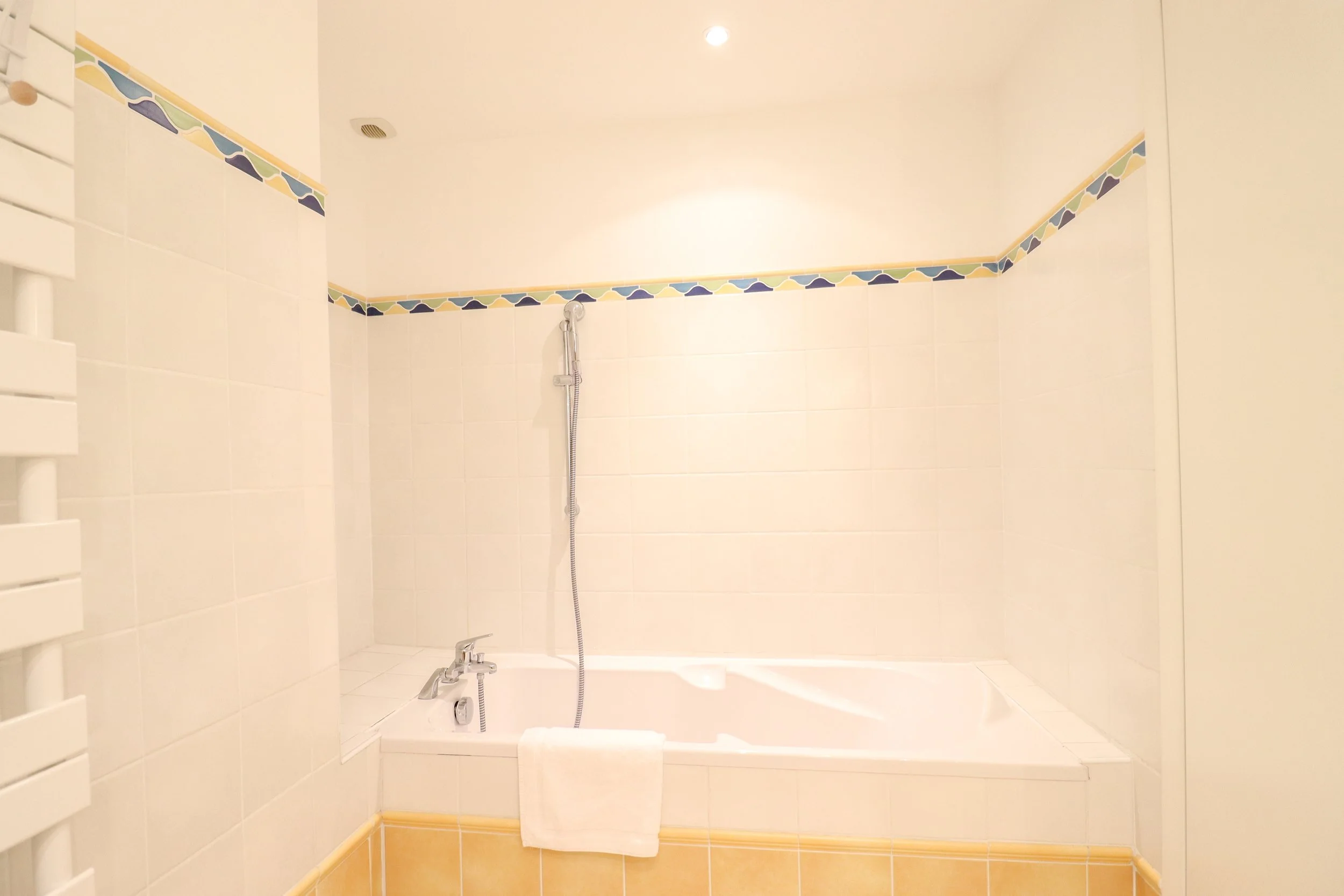 Salle de bain avec baignoire, têtière blanche, mur blanc avec une bande de carreaux décoratifs, robinet, douchette, serviette blanche.