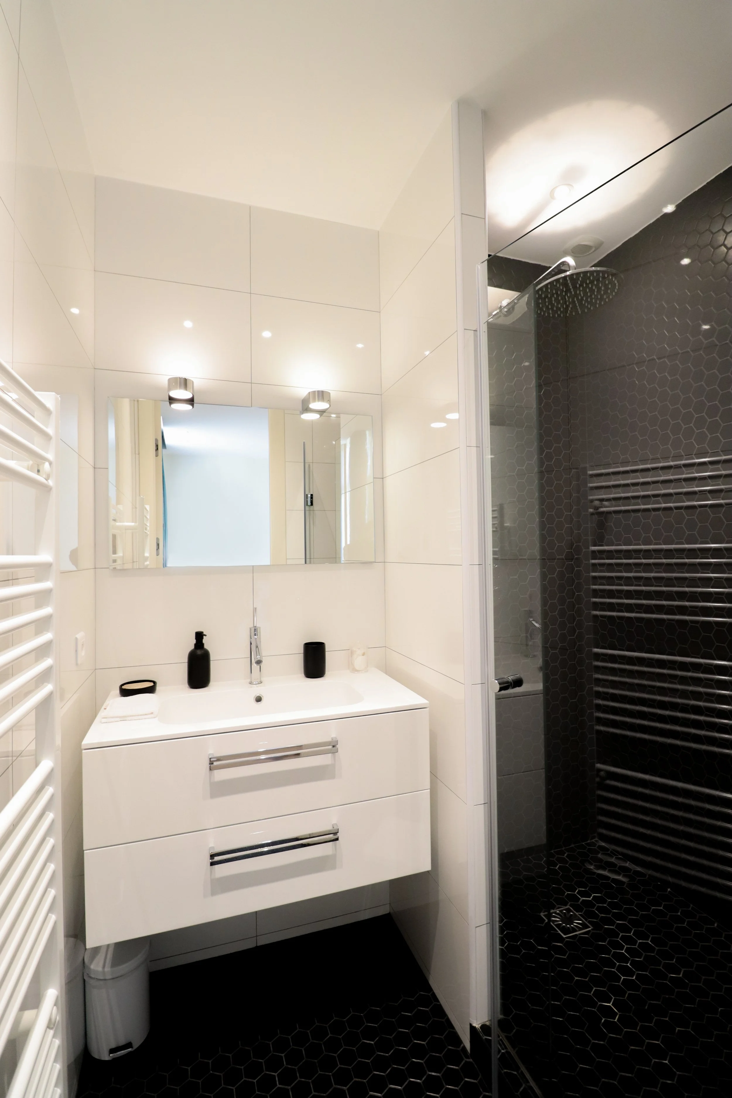 Salle de bain moderne avec lavabo blanc, miroir, douche à l'italienne avec paroi vitrée, murs noirs avec carrelage hexagonal, et radiateur torche