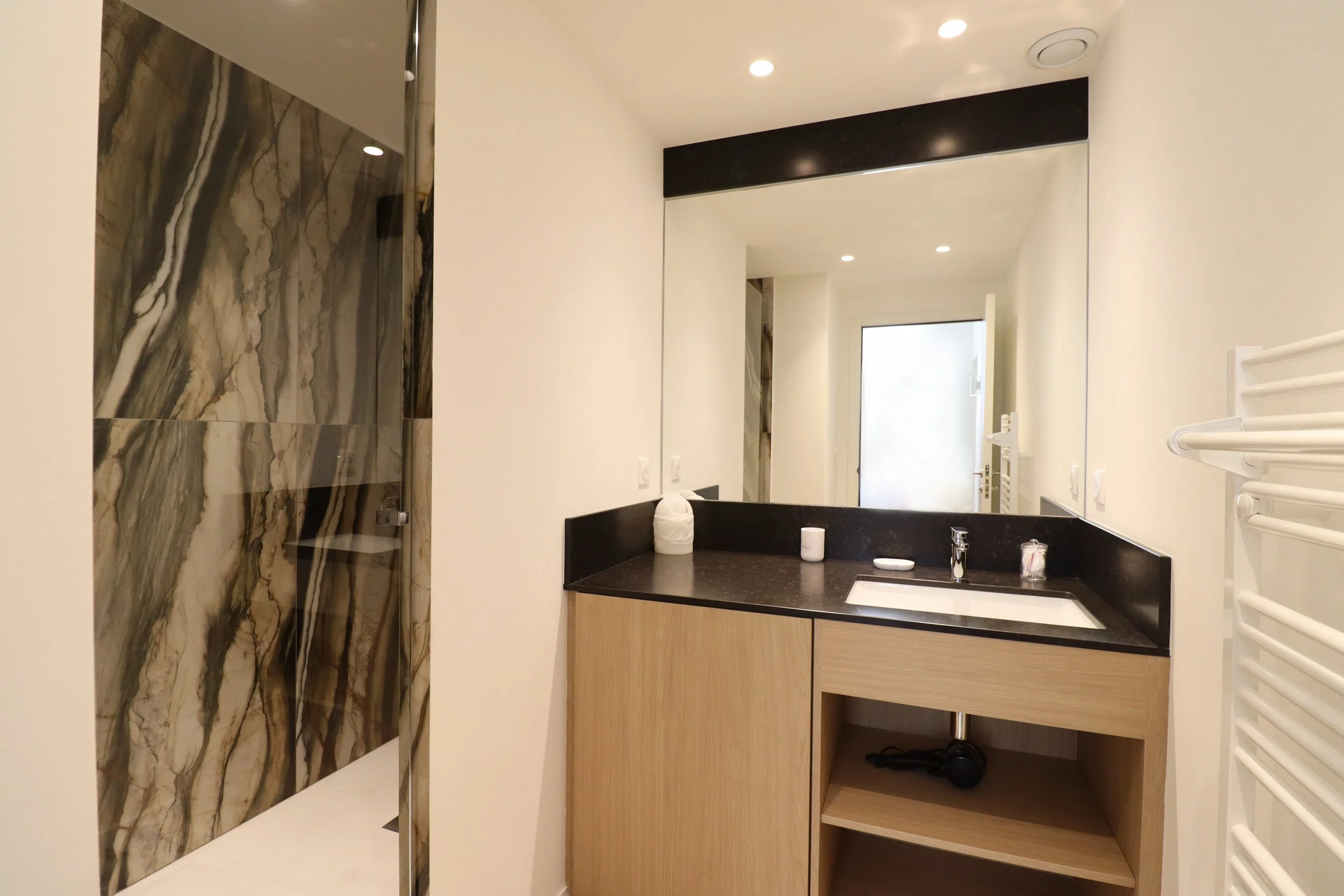 Vue d'une salle de bain moderne avec un miroir, un lavabo encastré dans un meuble en bois clair, et un mur en marbre avec un motif brun, beige et noir. Il y a également un radiateur électrique blanc à droite.