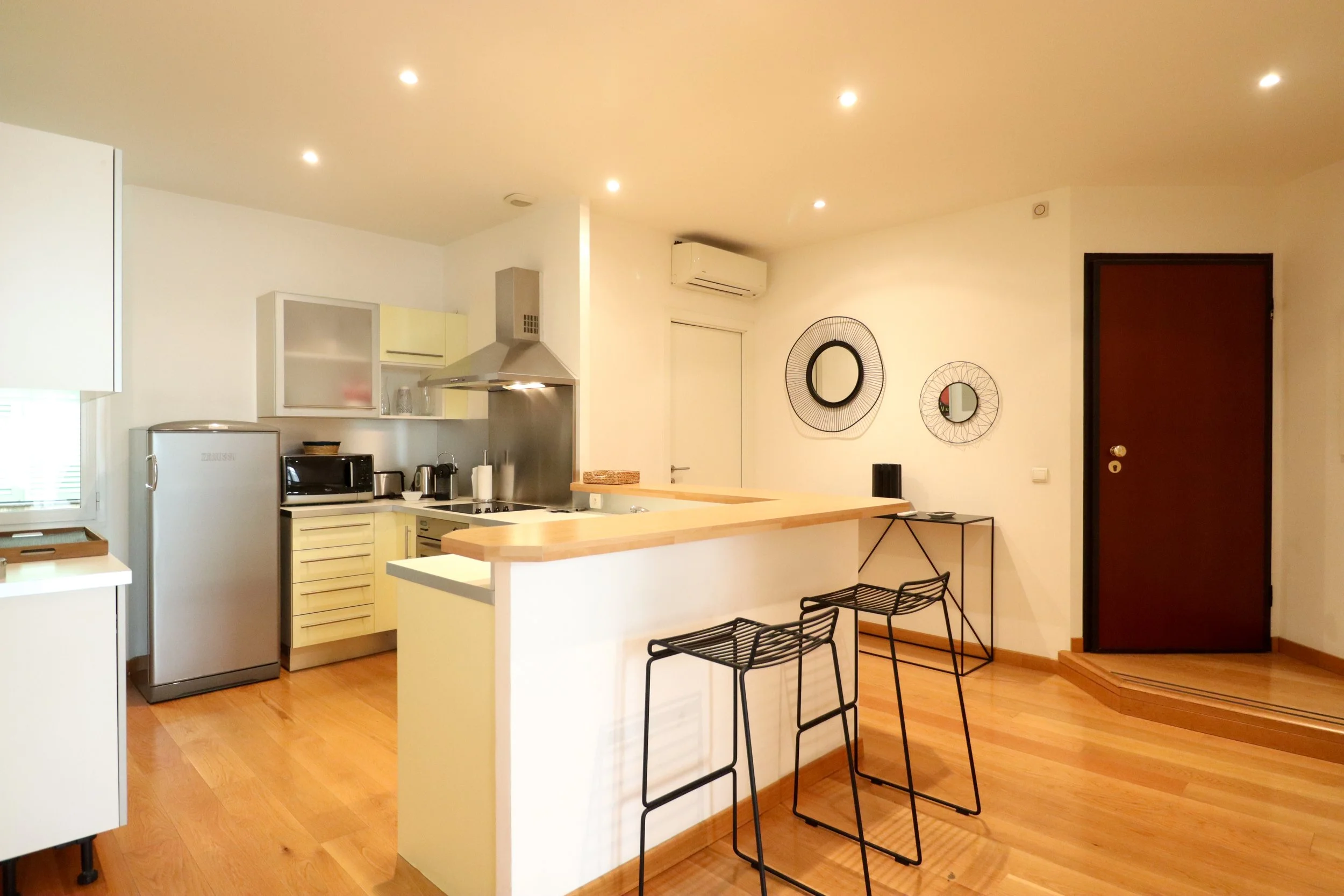 Cuisine avec bar en bois, chaises métalliques, réfrigérateur, four à micro-ondes, et décoration moderne avec deux miroirs ronds