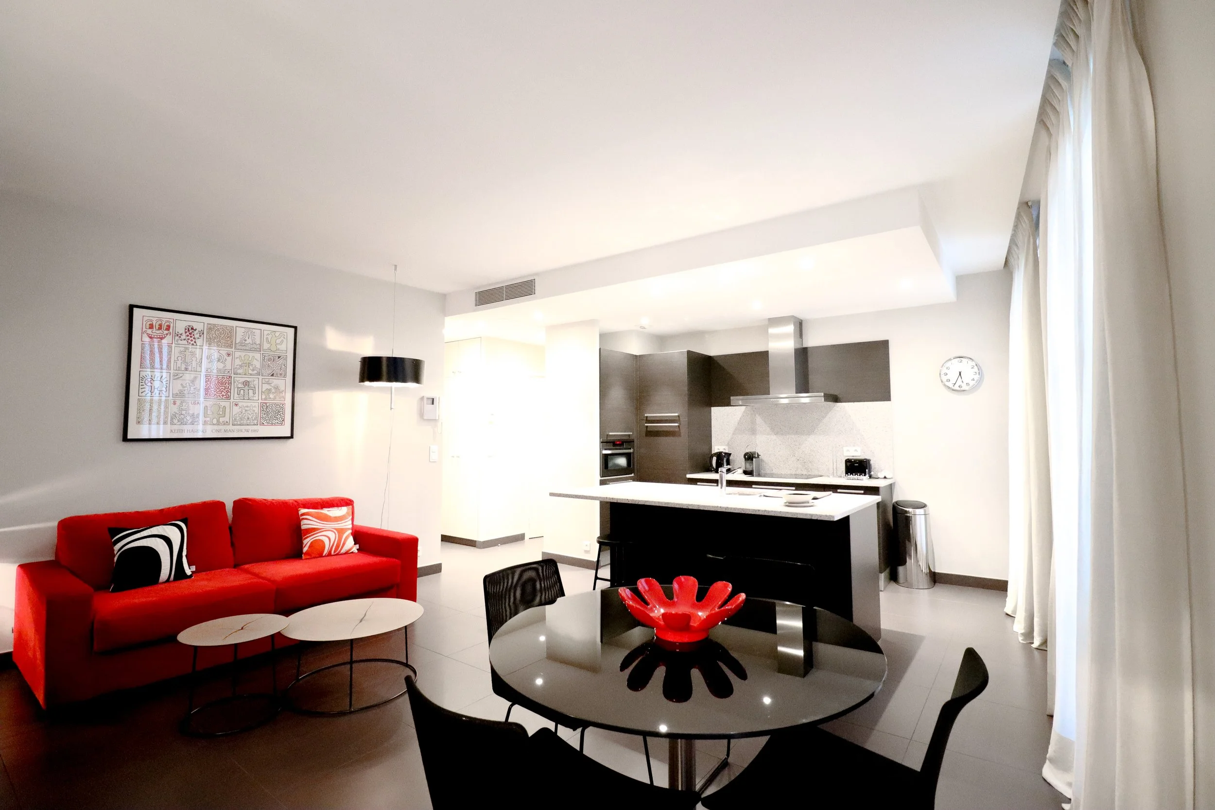 Intérieur moderne avec un canapé rouge, une table basse ronde, une cuisine en arrière-plan, et une horloge sur le mur. La pièce est lumineuse avec des grands rideaux blancs.
