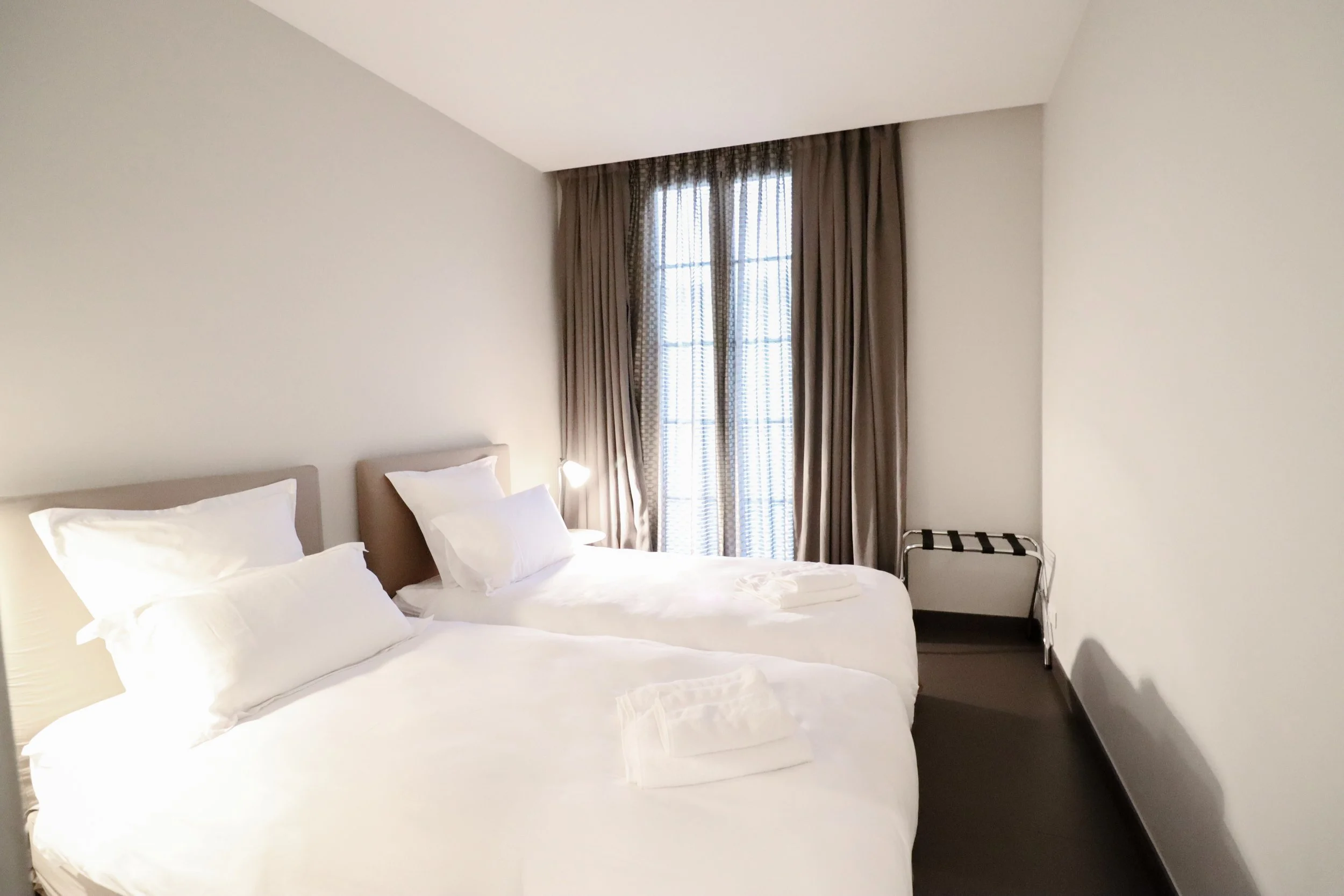Chambre avec deux lits maisons, fenêtres avec rideaux marron, lampe de chevet, panier à linge, murs blancs, sol sombre.