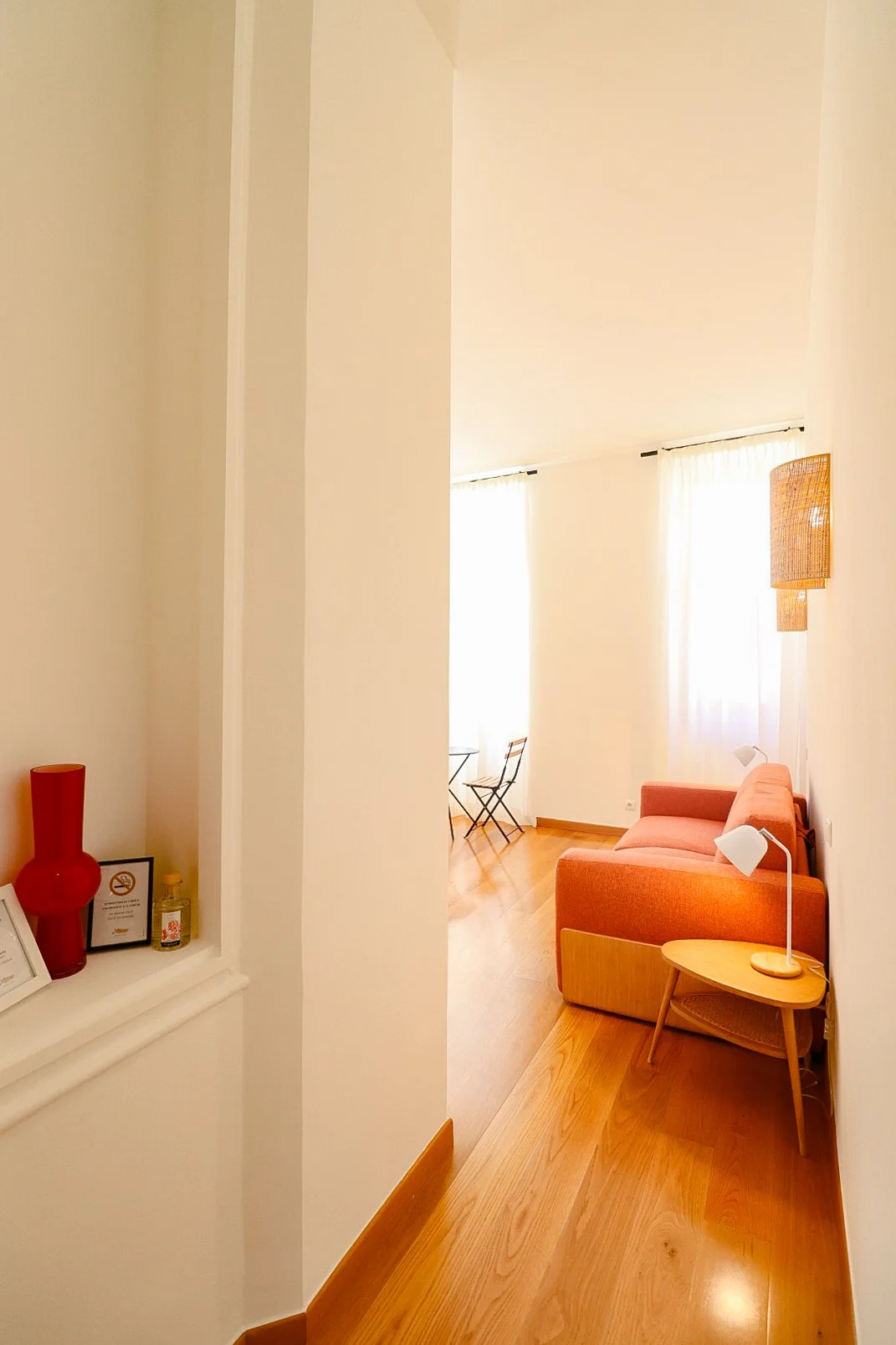 Salon lumineux avec un canapé rose, deux lampes de lecture blanches, une table basse en bois, des rideaux blancs, et deux chaises pliantes près d'une fenêtre.