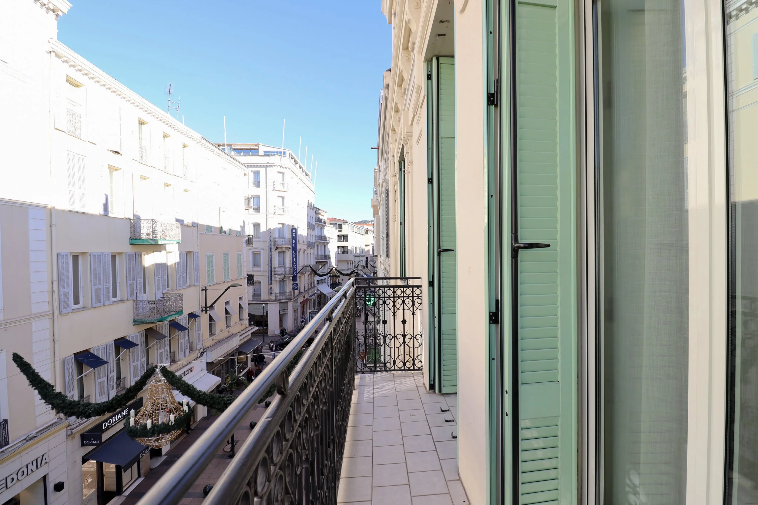 Vue depuis un balcon d'un bâtiment résidentiel montrant une rue commerçante animée avec plusieurs boutiques, des décorations de Noël et un ciel clair.