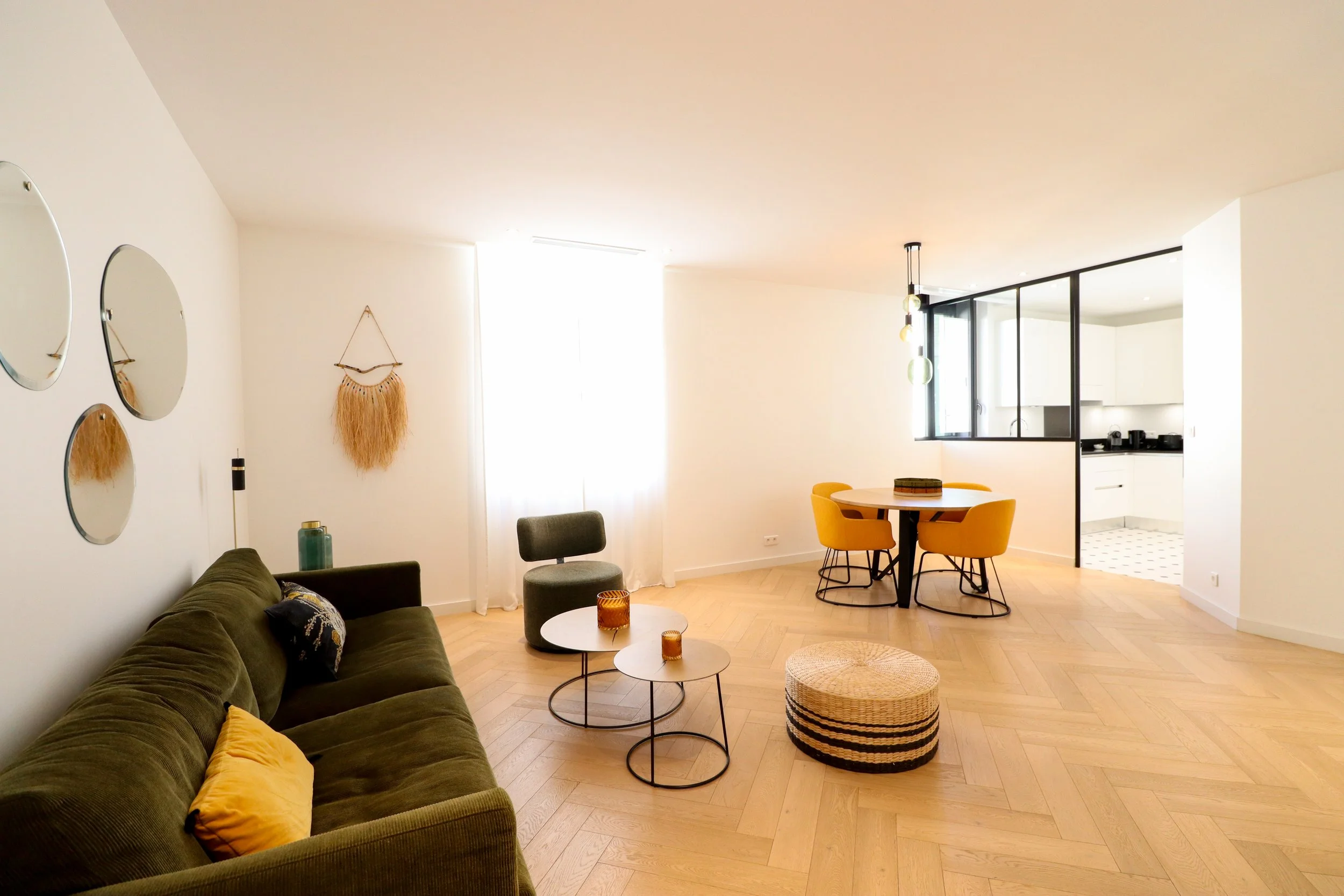Salon moderne avec un canapé vert, une table basse ronde, un fauteuil vert, et des décorations murales, dans un espace lumineux avec buffet blanc et cuisine moderne en arrière-plan.