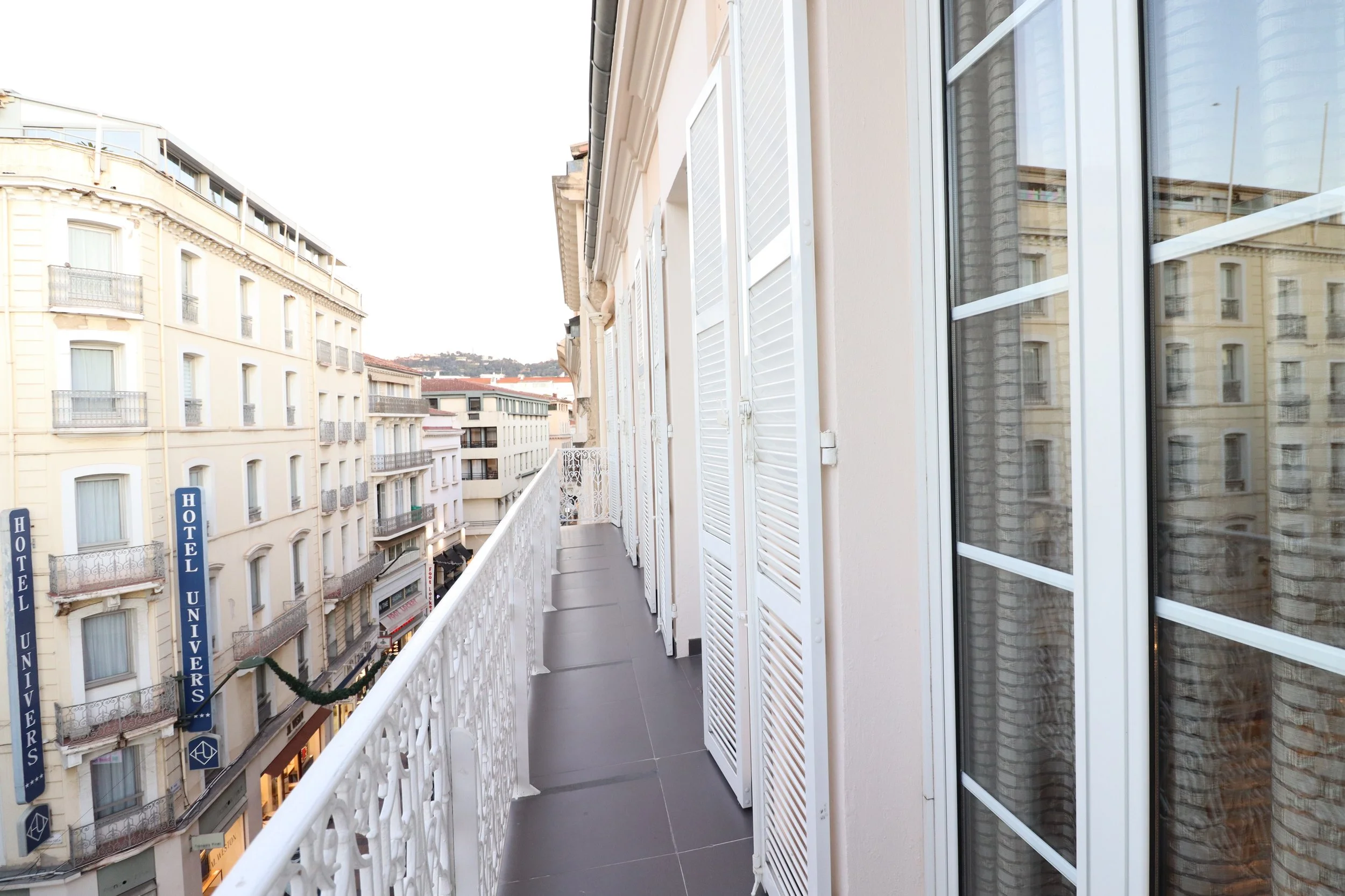 Balcon d'un appartement avec vue sur la ville, fermé par des volets blancs, avec des bâtiments d'hôtels et des enseignes commerce en ville.