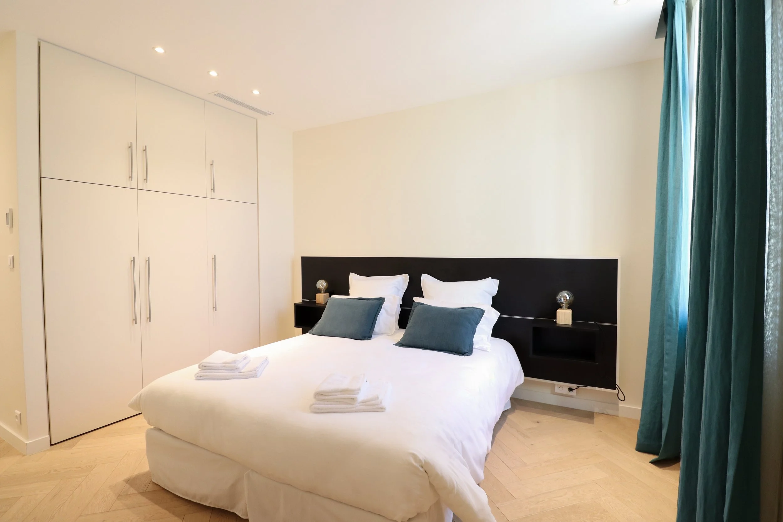 Chambre avec lit double, draps blancs, quatre serviettes pliées sur le lit, deux coussins gris, une tête de lit noire, deux lampes de chevet avec lampes en forme de sphère, un grand placard blanc avec poignées métalliques, mur blanc, fenêtre avec rid