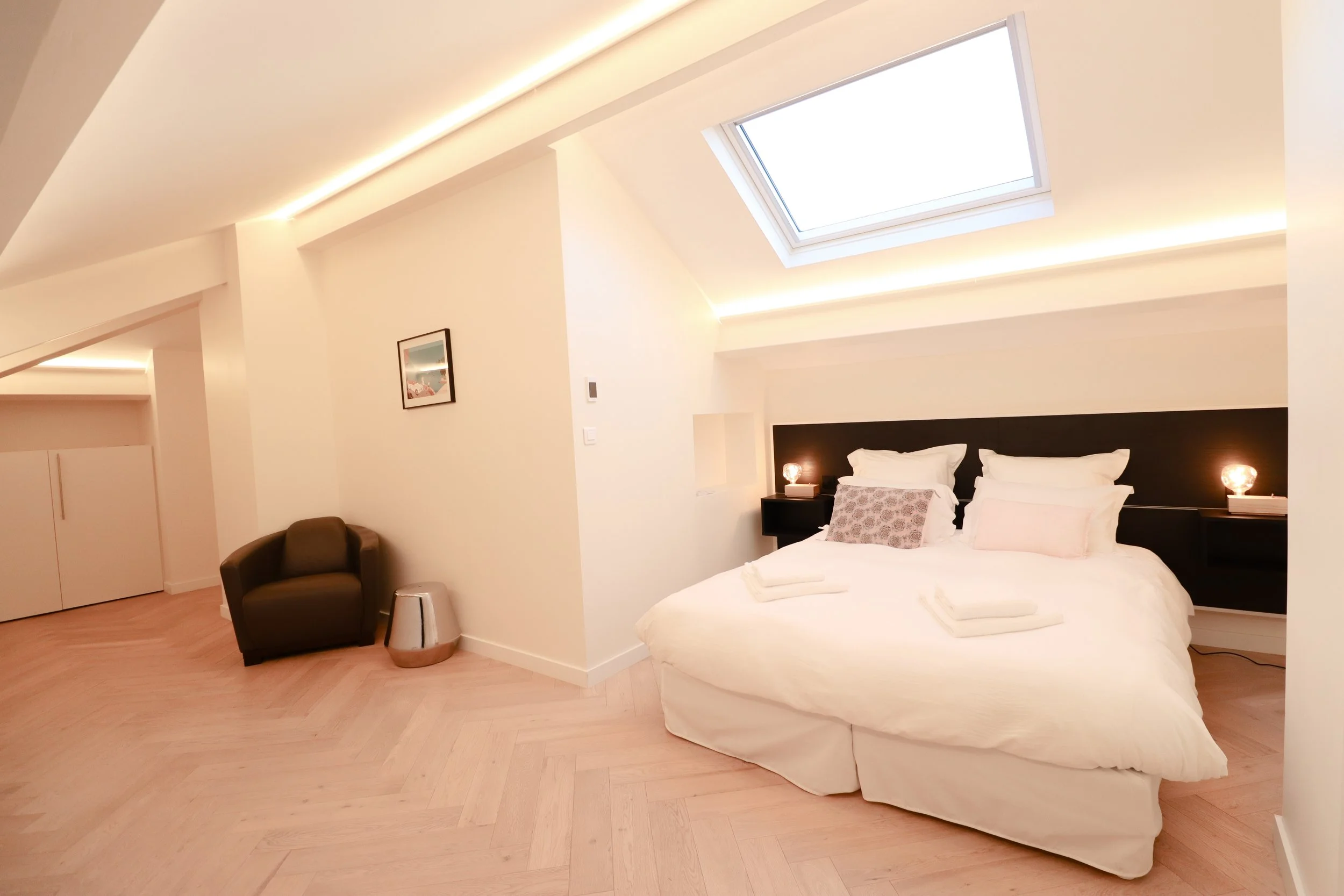 Chambre moderne avec un lit king-size, deux lampes de lecture, un fauteuil noir, une poutre avec un plafond en pente et un plafond avec un puits de lumière.