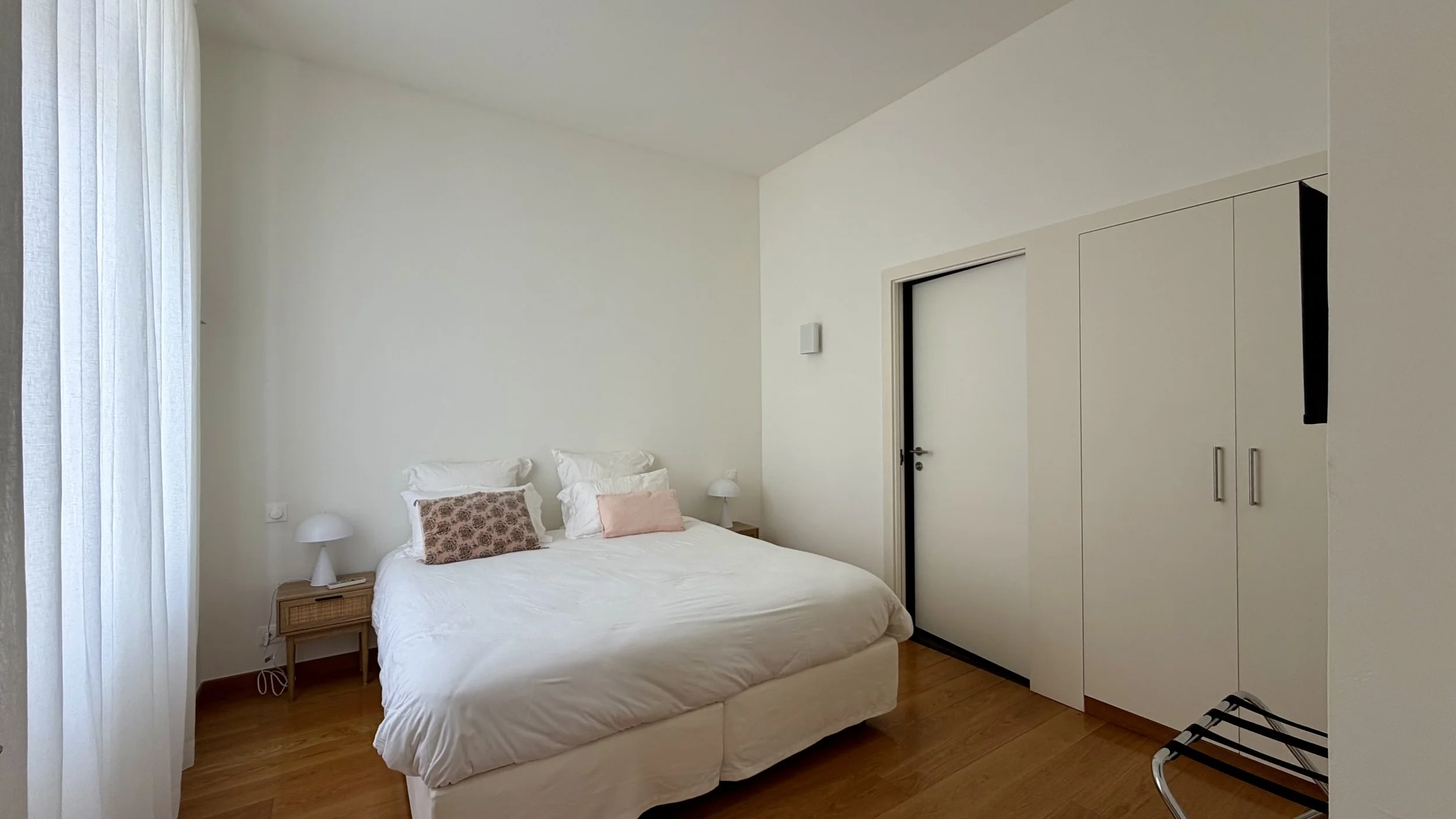 Chambre minimaliste avec un lit blanc, deux oreillers, une petite table de nuit avec une lampe, un meuble avec des portes blanches, une chaise pliante et une fenêtre avec rideaux blancs.