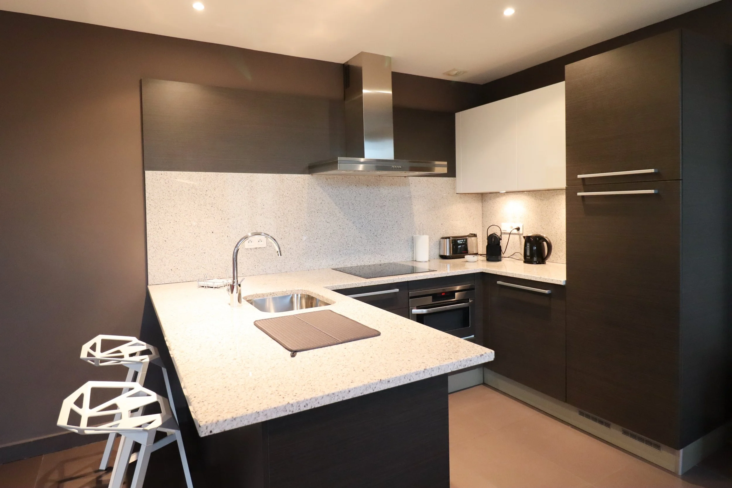 Cuisine moderne avec comptoir en quartz, four encastré, hotte en métal, et ustensiles de cuisine noirs, avec chaises blanches à motifs