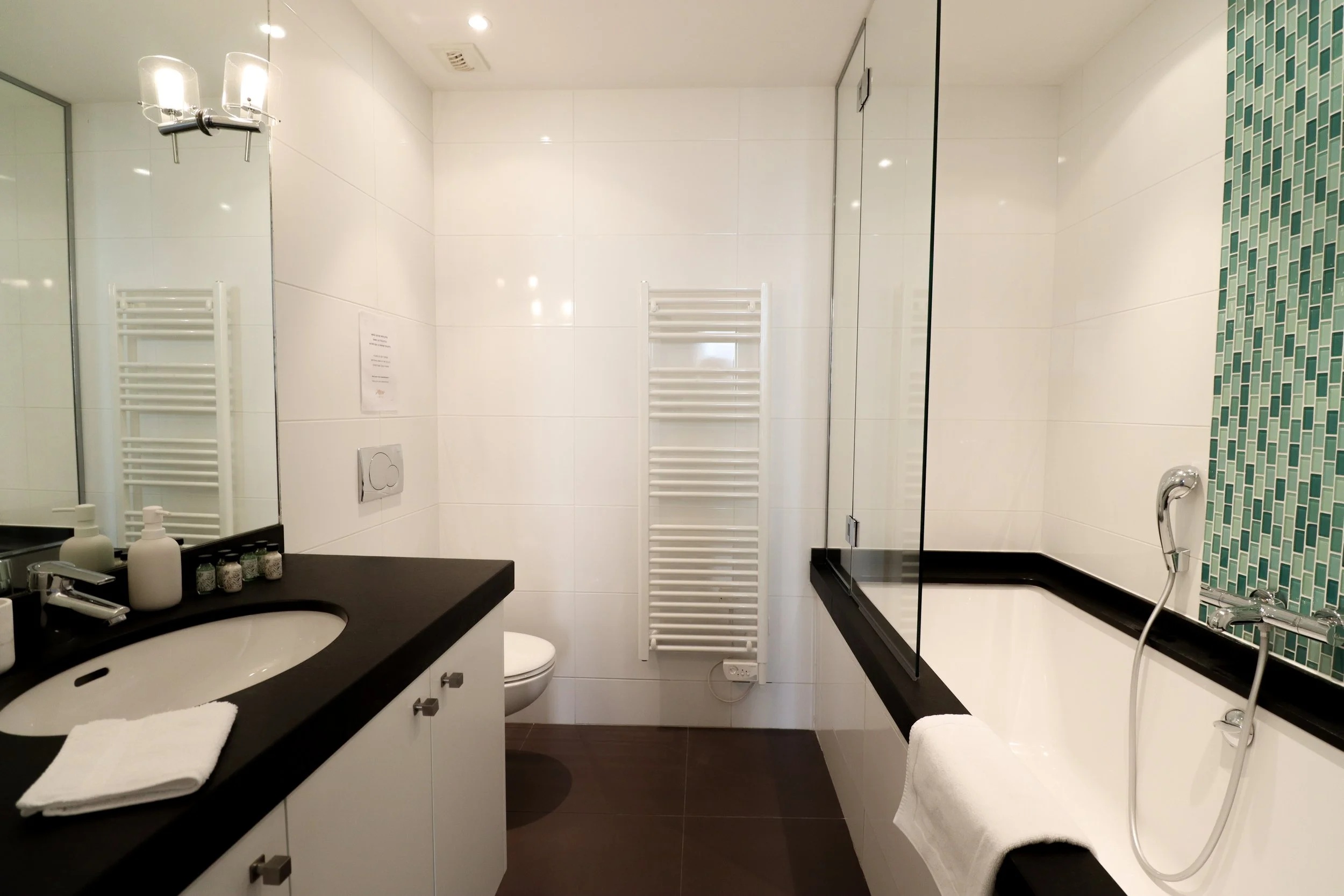 Salle de bain moderne avec miroir, lavabo, baignoire, douche, sèche-serviettes blanc, accessoires de toilette, décor épuré et sol en bois foncé.