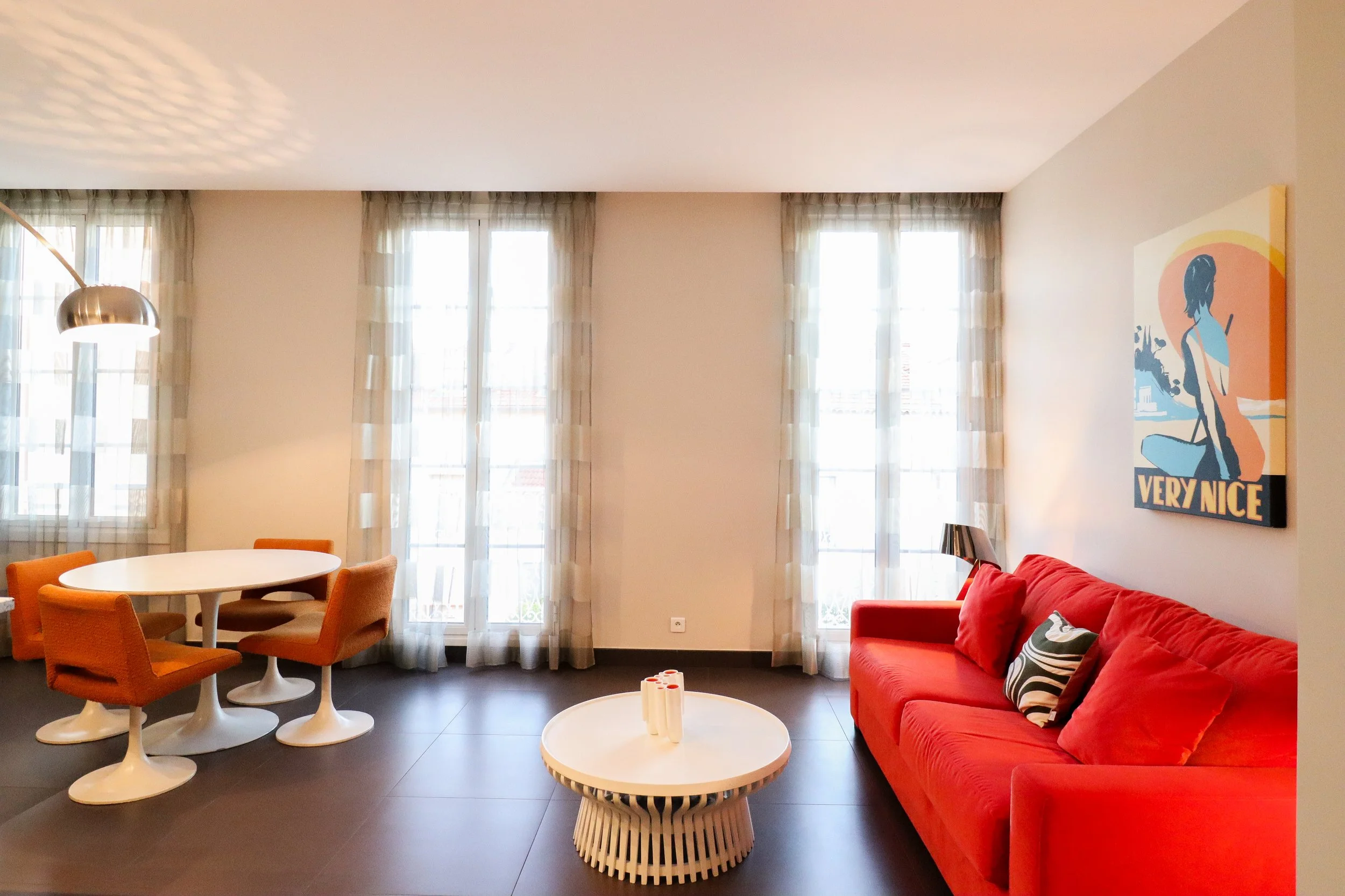 Salon lumineux avec trois grandes fenêtres, un canapé rouge, une table ronde blanche, des chaises orange, une lampe suspendue en métal, une peinture moderne représentant une femme avec une fleur, et un décor moderne
