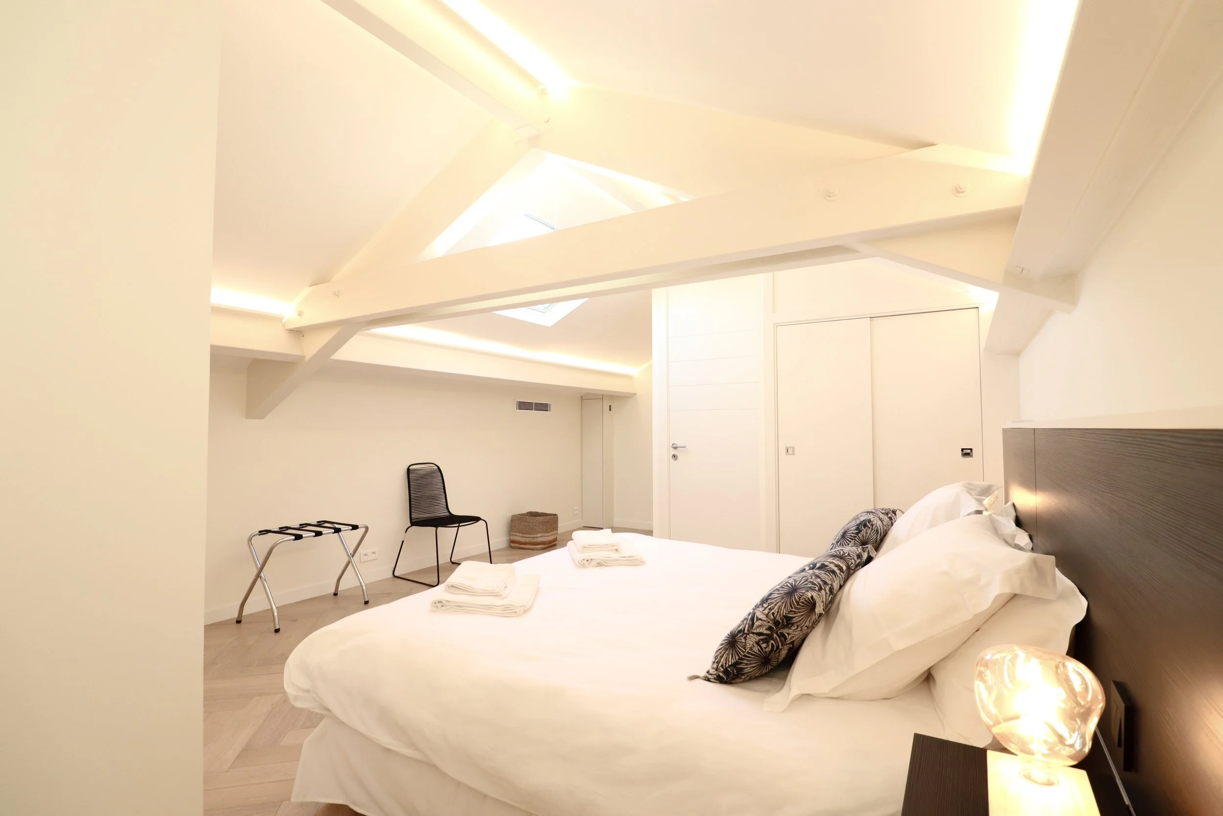  chambre avec lit blanc, oreillers, lampes de chevet, chaise, porte, plafond avec lumières encastrées, fenêtres de toit.