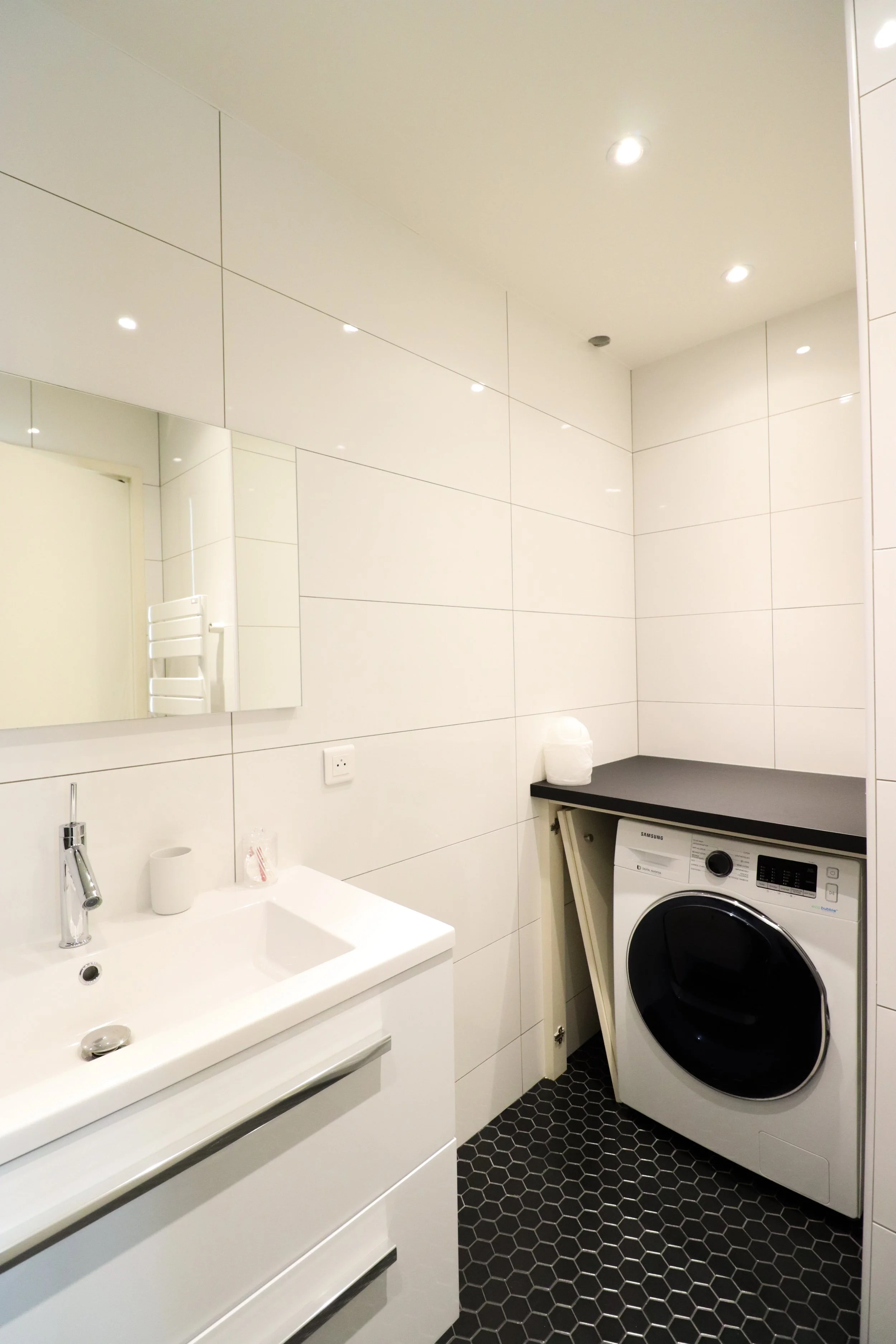 Salle de bain avec une vasque blanche, un lavabo, un miroir, un sèche-serviettes blanc, une machine à laver, et un comptoir noir, avec des murs et un plafond blancs et un sol en carreaux hexagonaux noirs.