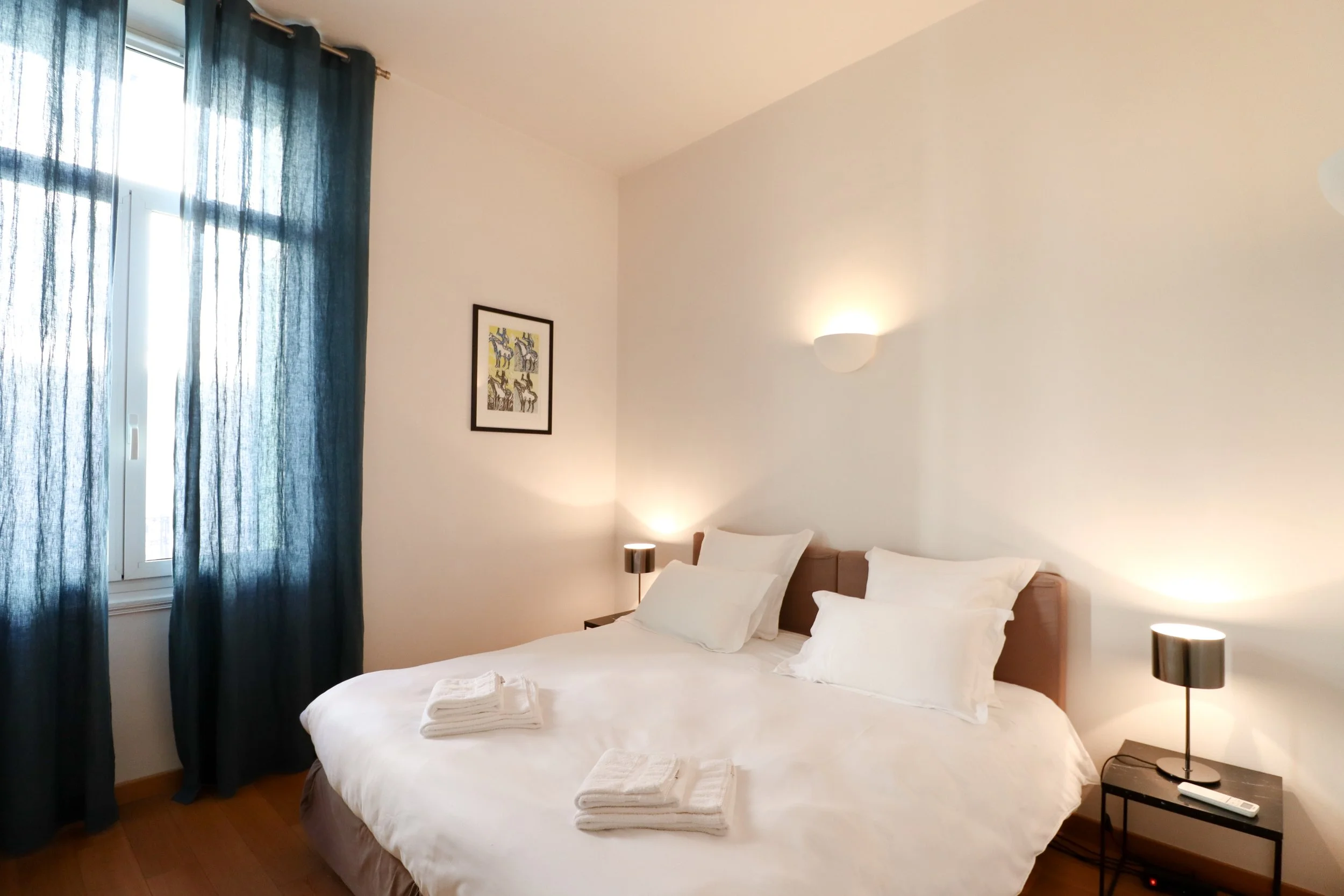 Chambre avec lit double, draps blancs, serviettes pliées sur le lit, fenêtre avec rideaux bleus, tableau avec silhouettes d'outre-toi, lampes de chevet, face au lit et murs blancs.