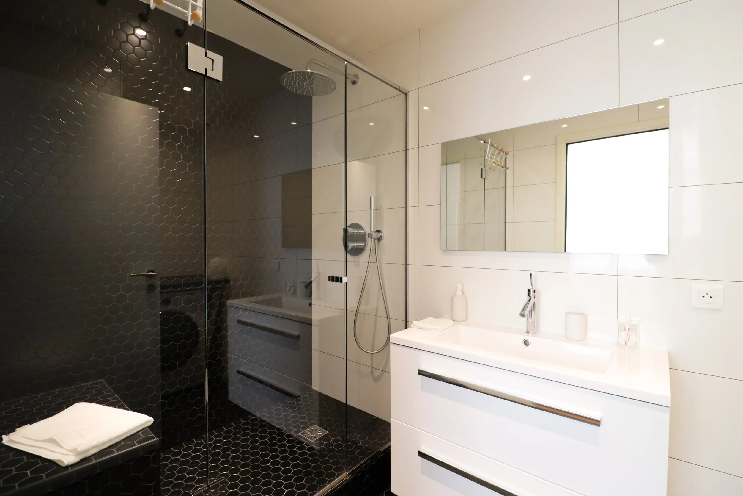 Salle de bain moderne avec douche à l'italienne en verre, carrelage noir hexagonal, meuble blanc avec miroir, lavabo avec robinets, et fenêtres laissant entrer la lumière naturelle.
