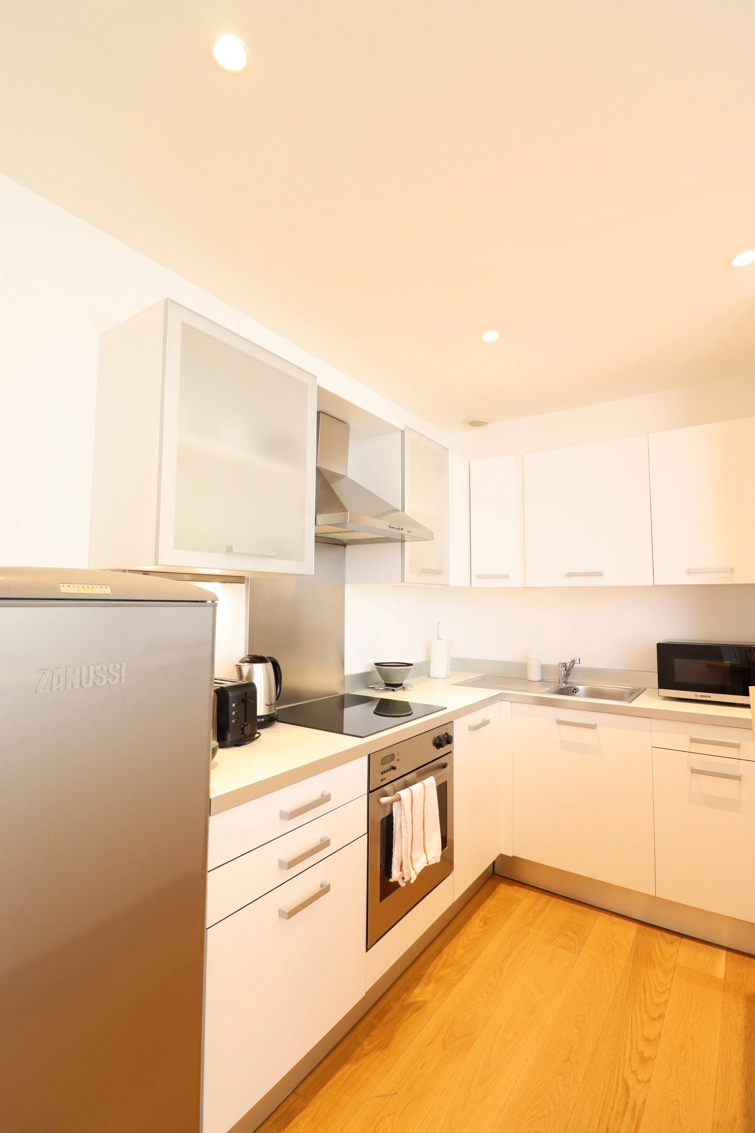 Cuisine moderne blanche avec électroménagers en acier inoxydable, comptoir avec petits appareils, tiroirs et placards, plafond avec éclairages encastrés, sol en bois clair.