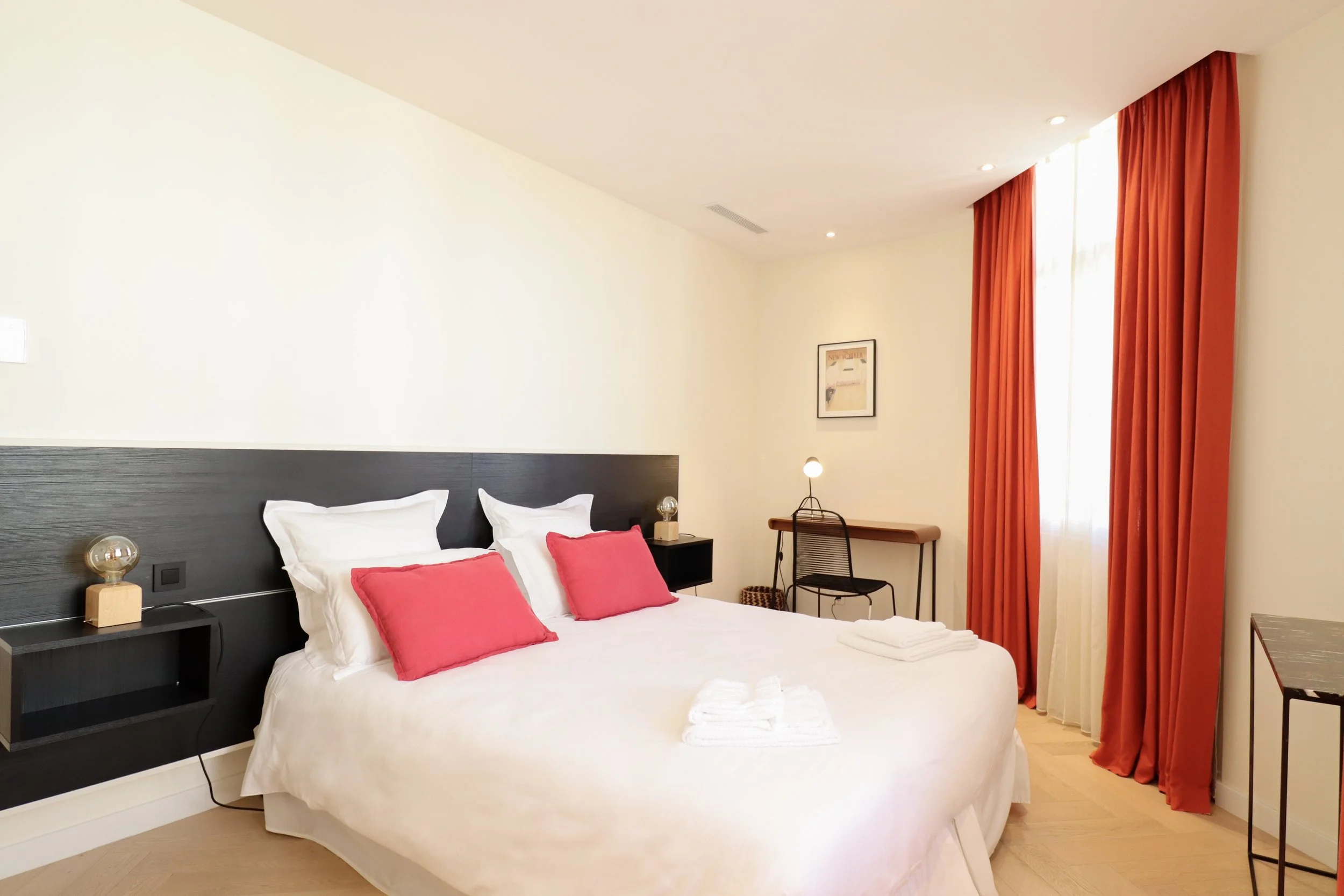 Chambre d'hôtel moderne avec lit à linge blanc et coussins roses, grande fenêtre avec rideaux orange, bureau en bois et chaise noire, décor minimaliste et éclairage chaud.