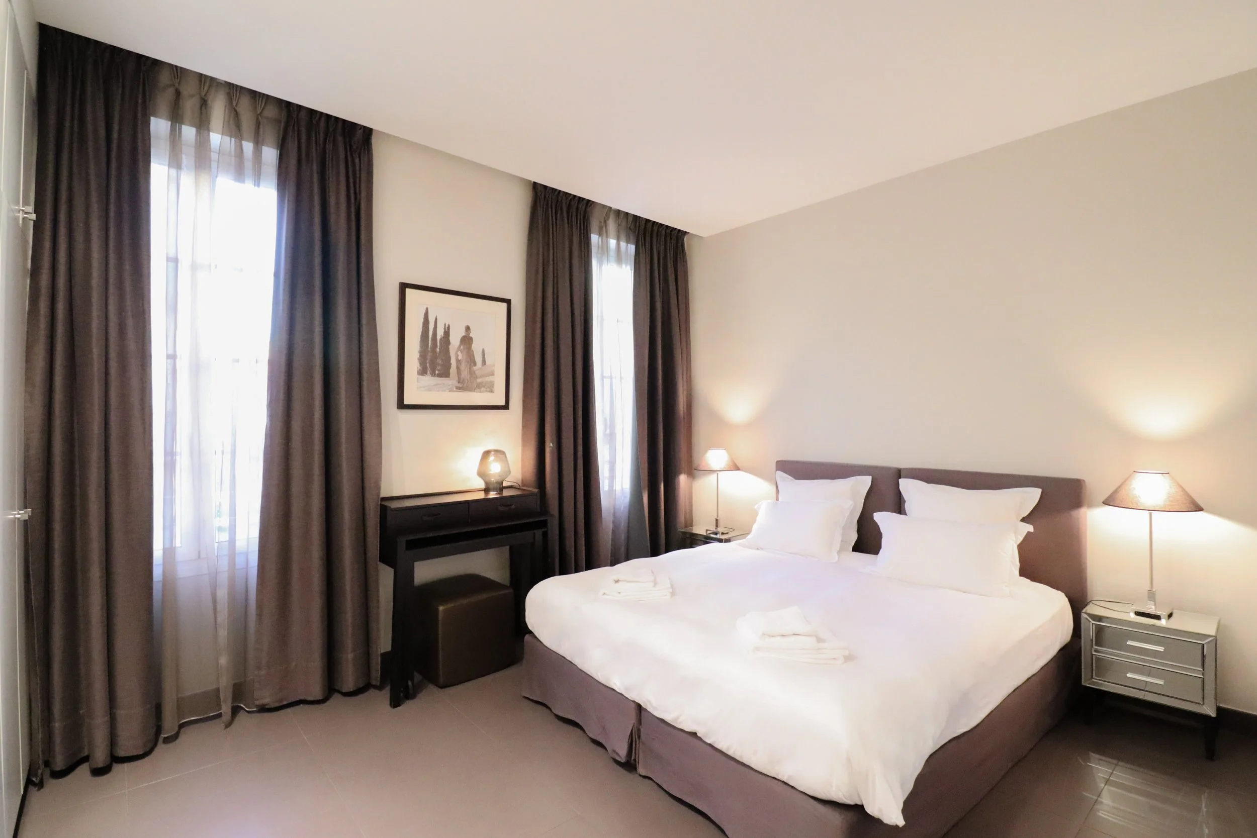 Chambre d'hôtel avec lit double, deux lampes de chevet, rideaux sombres, photo encadrée au mur, et meubles noirs.