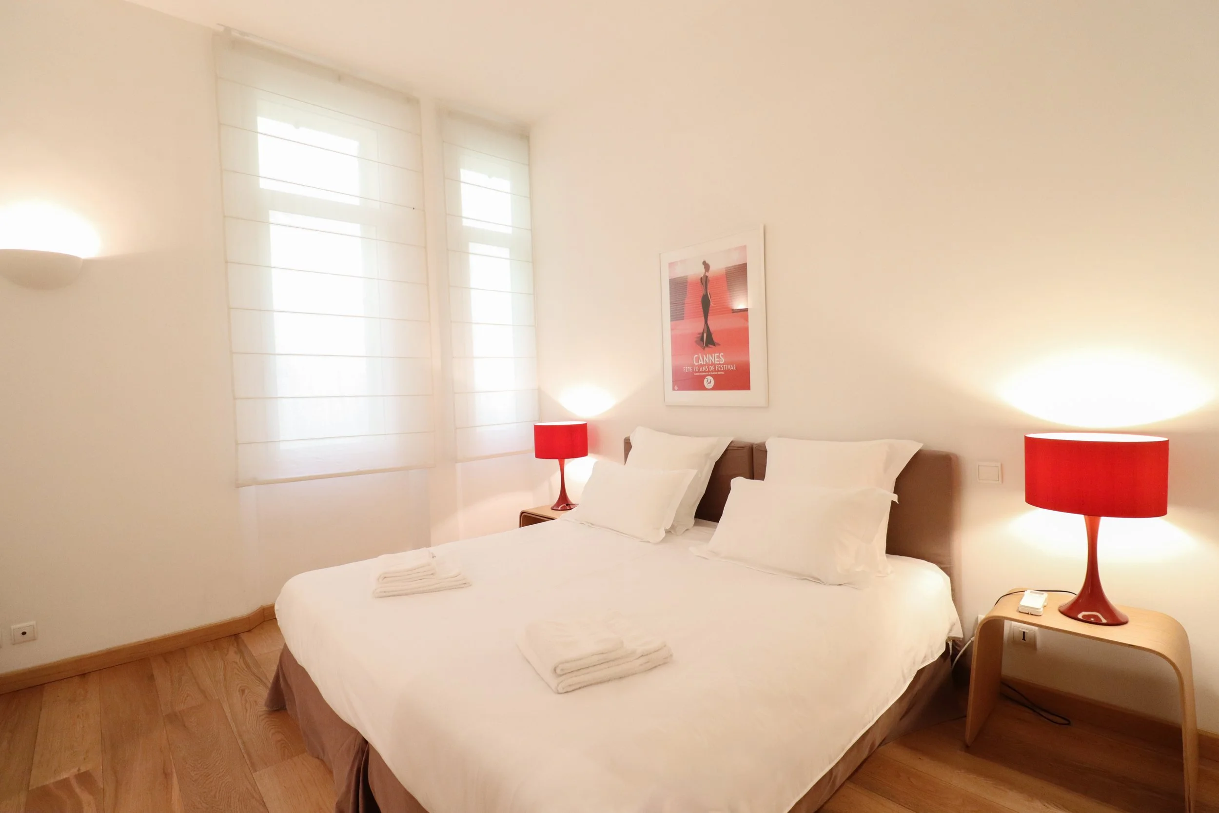 Chambre avec lit double, deux lampes rouges de chaque côté du lit, poster du festival de Cannes sur le mur, fenêtres avec volets blancs, sol en bois clair.