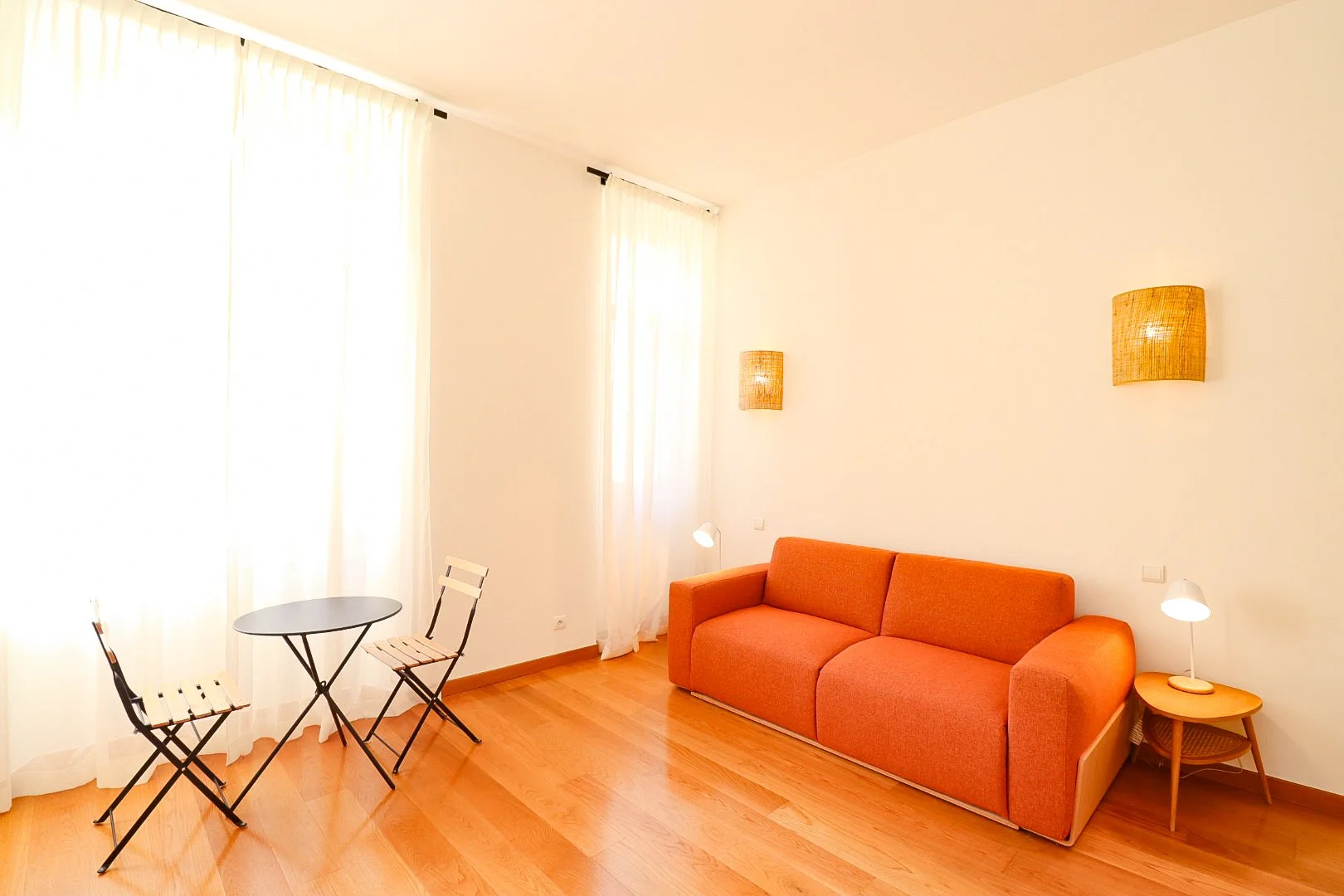 Salon avec canapé orange, deux lampes à côté, une petite table ronde avec deux chaises, rideaux blancs, murs blancs, plancher en bois