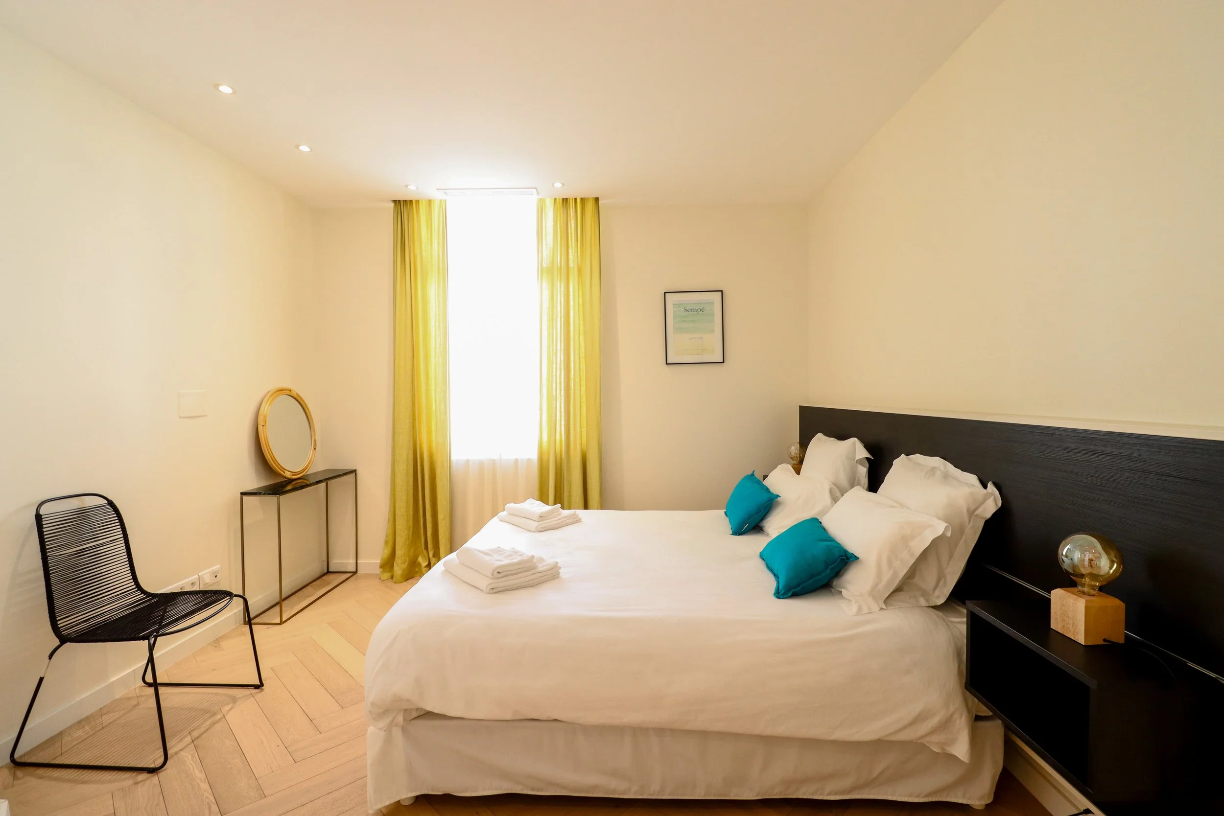 Chambre d'hôtel avec un lit double, des oreillers bleus, lampes de chevet, un mur blanc, une fenêtre avec rideaux jaunes, un miroir rond sur une table, une chaise noire en métal, un tableau au mur, sols en bois clair.
