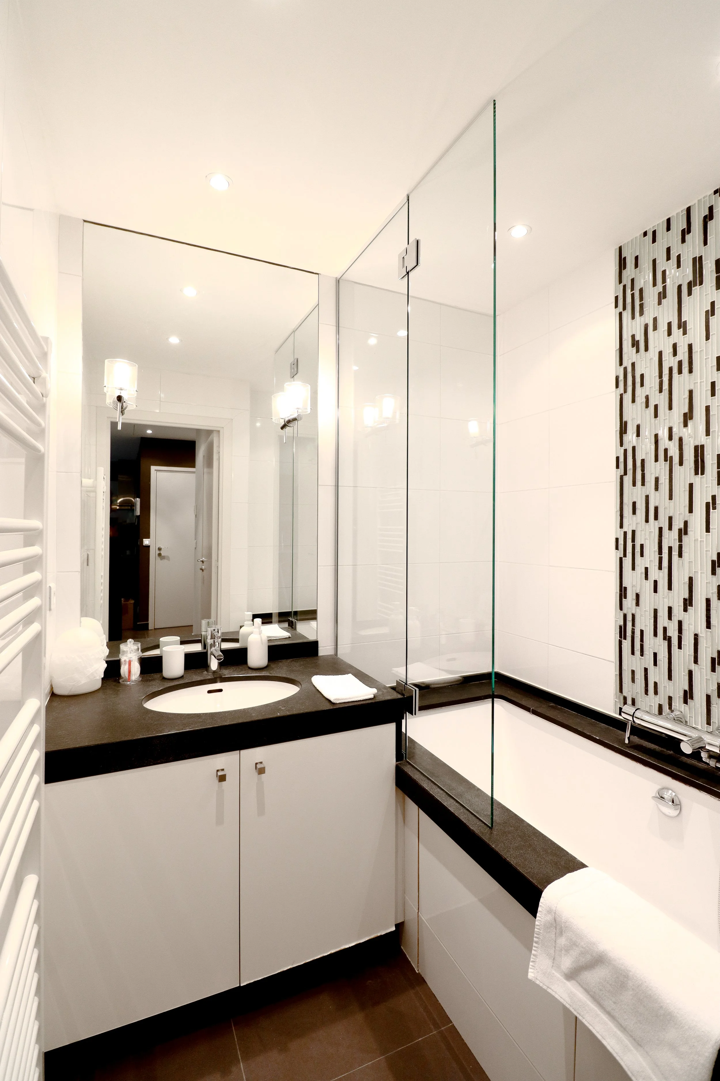 Salle de bain moderne avec miroir, lavabo, comptoir noir, douche avec porte en verre, mur en carreaux blancs et motif de mosaïque, luminaires, serviettes blanches, surface propre et bien éclairée.