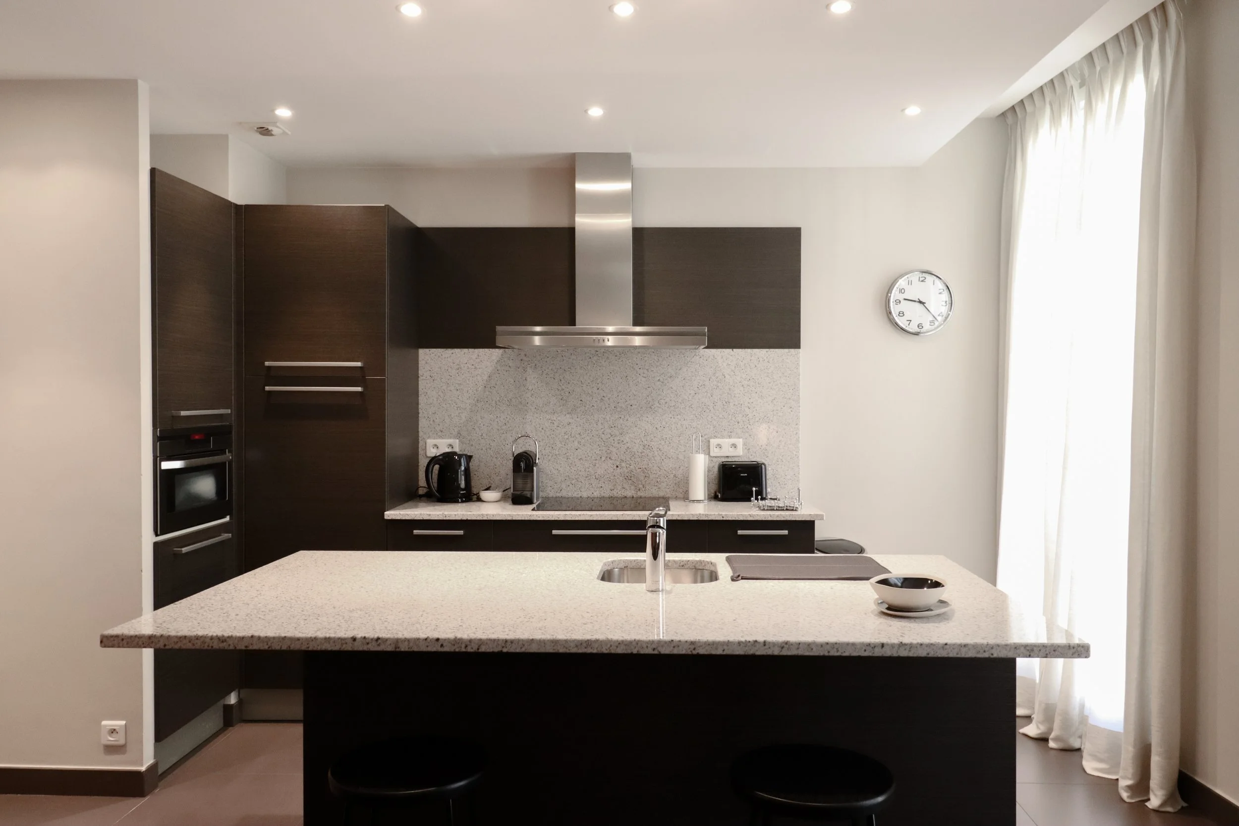 Cuisine moderne avec îlot central, comptoir en pierre, électroménagers intégrés, horloge murale, et fenêtres avec rideaux blancs.