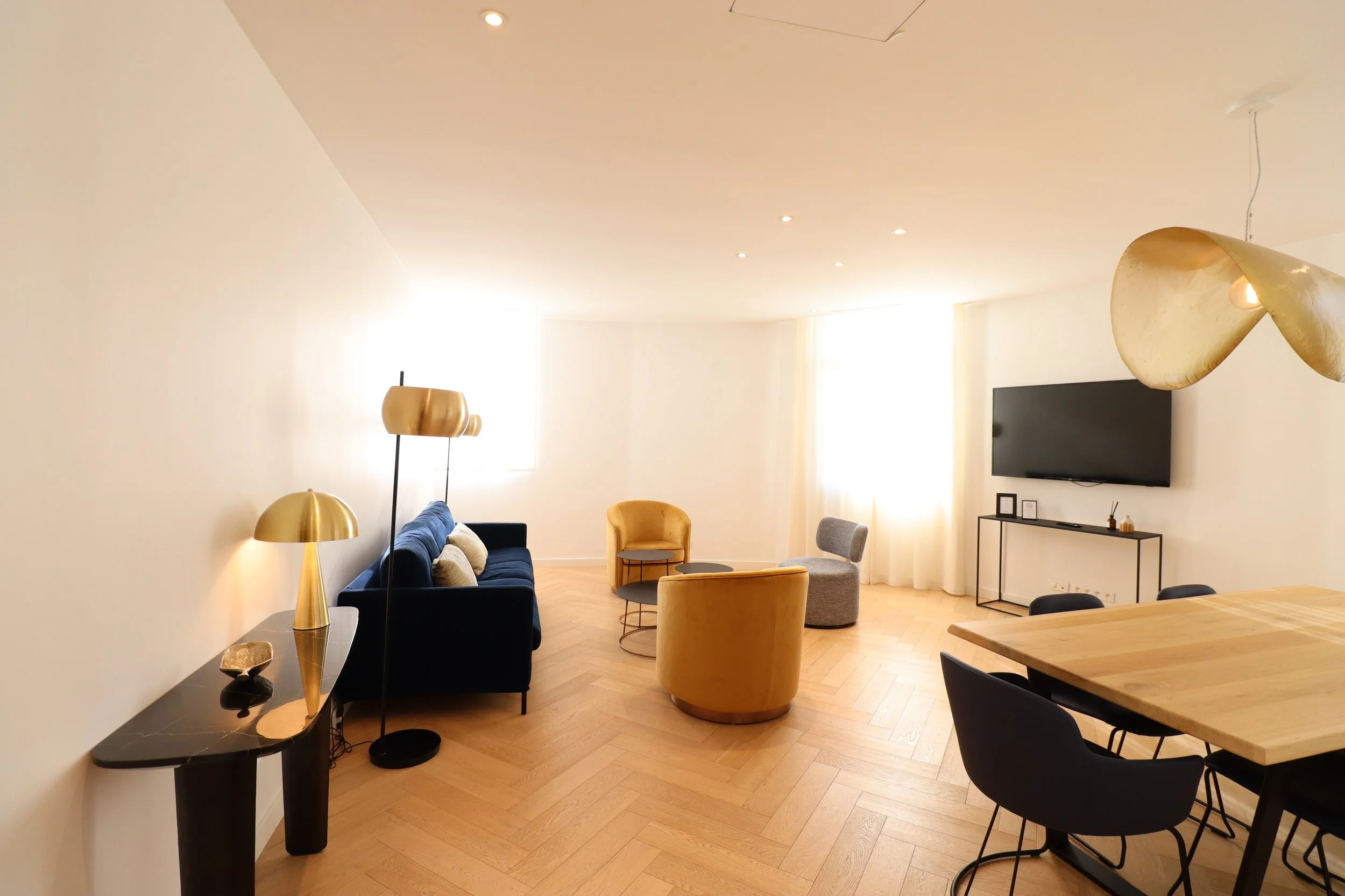 Salon moderne avec plusieurs fauteuils jaunes et gris, canapé bleu, tables basses, télévision murale, lampes en or, et table en bois, avec murs blancs et fenêtres lumineuses.