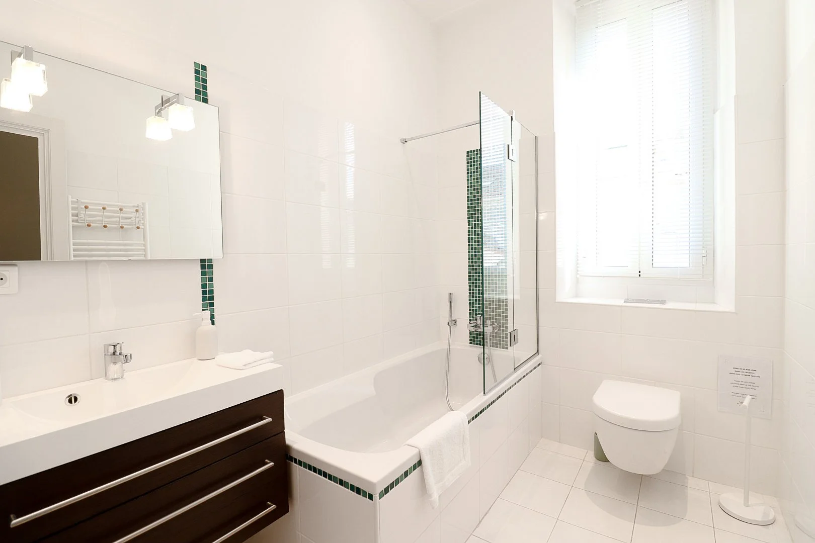 Salle de bain moderne avec une baignoire, un lavabo avec un miroir, un WC, une fenêtre à volets, et un rideau en verre séparant la baignoire.
