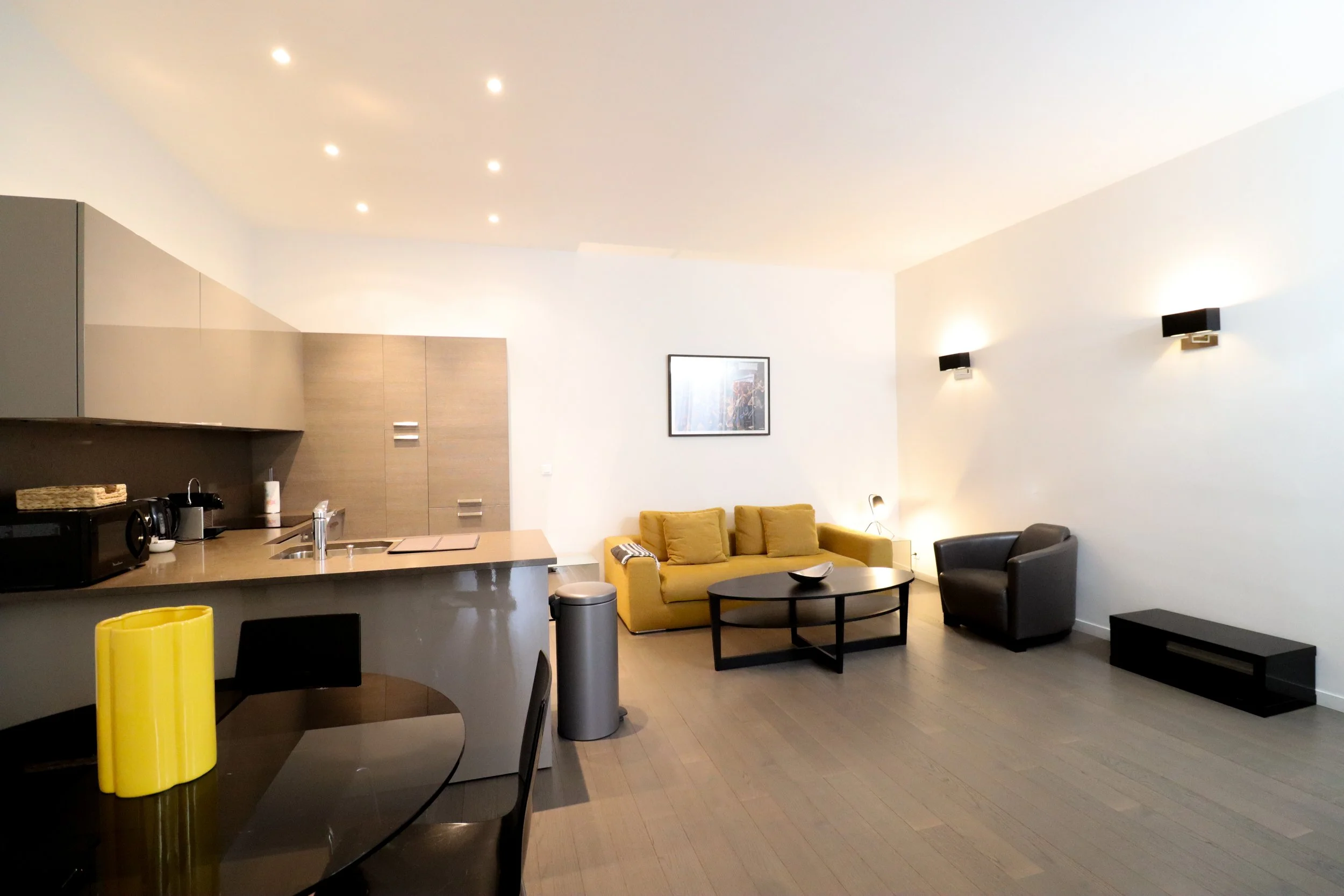 Intérieur d'un salon moderne avec un canapé jaune, deux fauteuils (un noir et un gris), une table basse noire, des murs blancs, des éclairages muraux, une peinture encadrée et un espace cuisine en arrière-plan.
