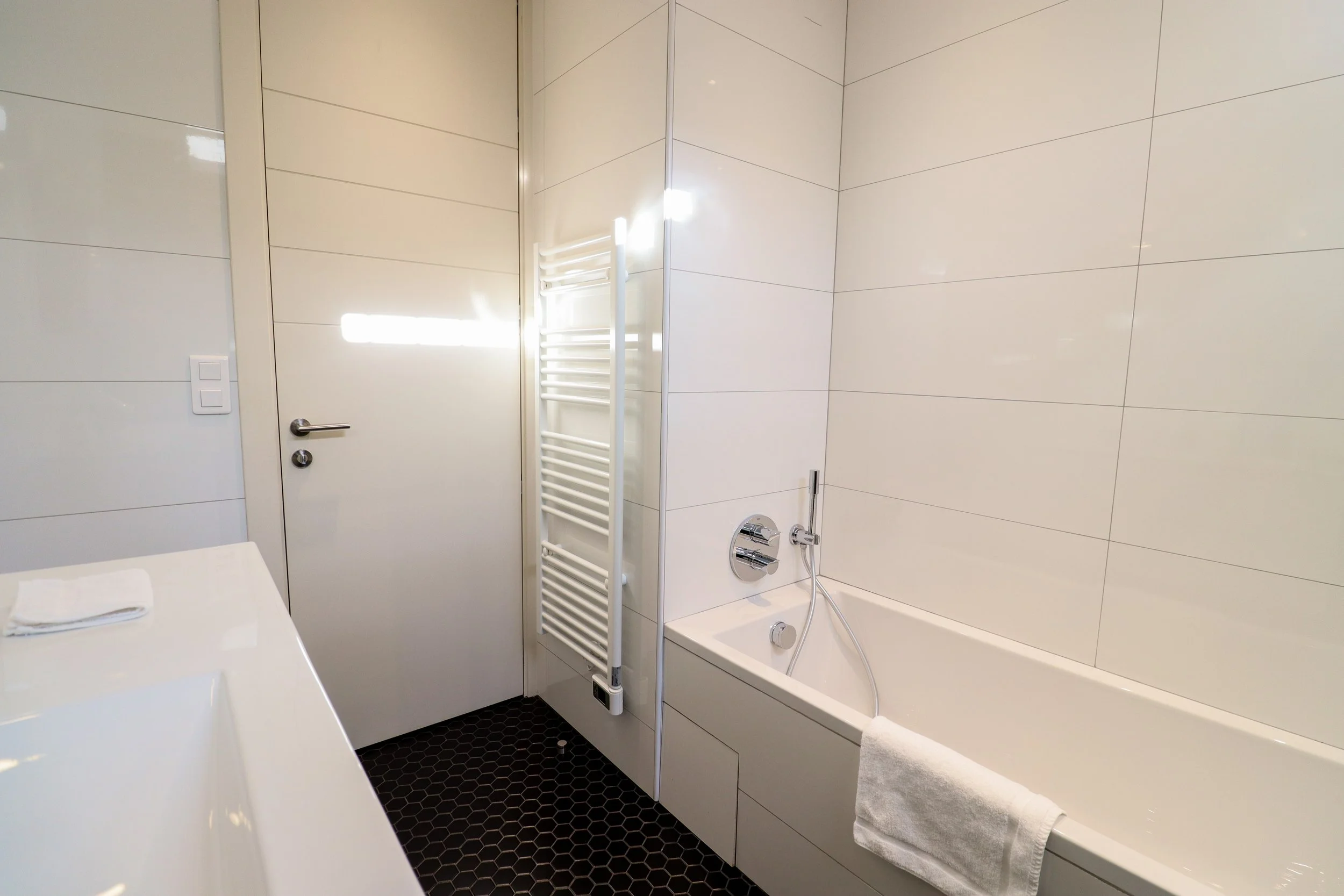 Salle de bain avec baignoire, mur blanc, porte blanche, radiateur blanc, robinetterie moderne, serviette blanche sur la baignoire, sol en carrelage noir hexagonal.