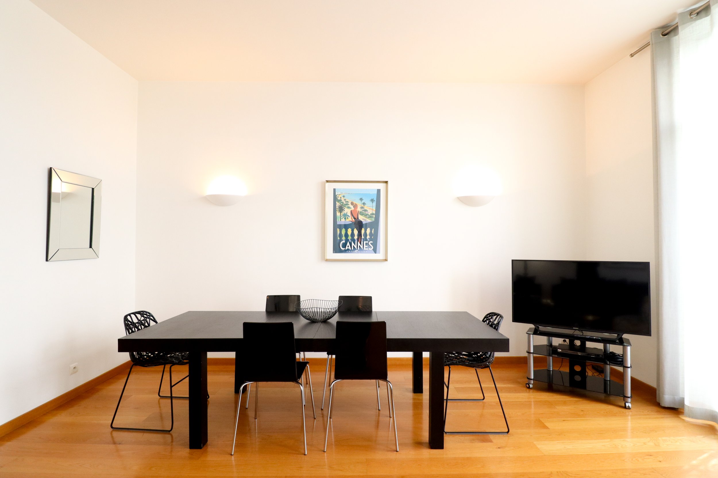 Salle à manger avec table noire, chaises en plastique noir, mur blanc, deux luminaires muraux, tableau avec slogan 'CANNES', télévision sur meuble en métal.