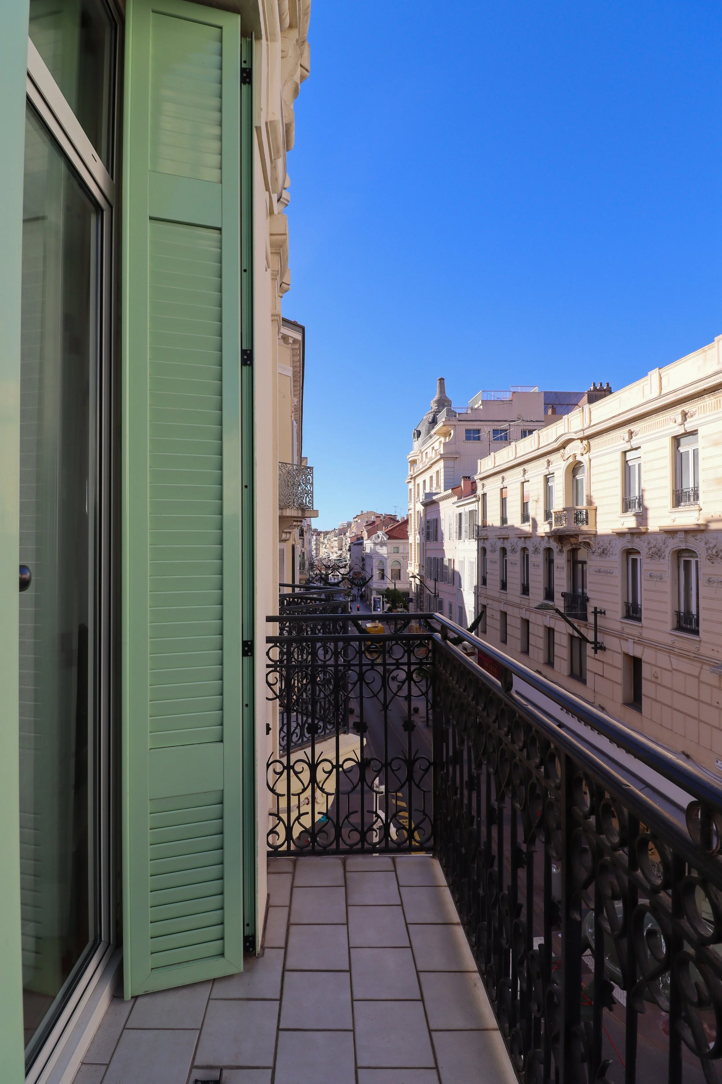 Balcon d'un appartement avec des portes fenêtres vertes et un rebord en métal noir, face à une rue bordée d'immeubles haussmanniens en pierre beige sous un ciel clair.