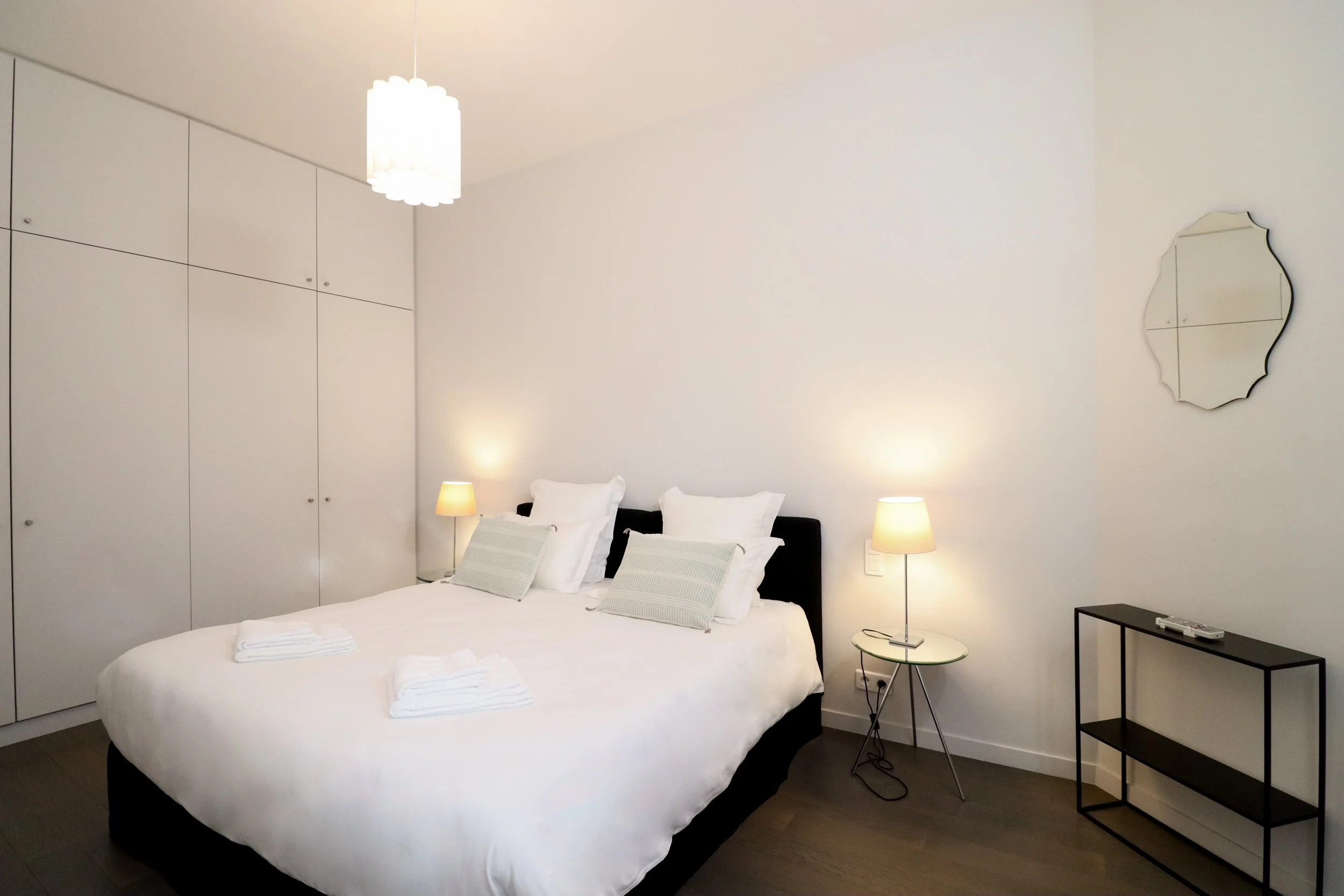 Chambre moderne avec lit double, deux tables de chevet avec lampes, mur blanc avec miroir décoratif et armoires intégrées.