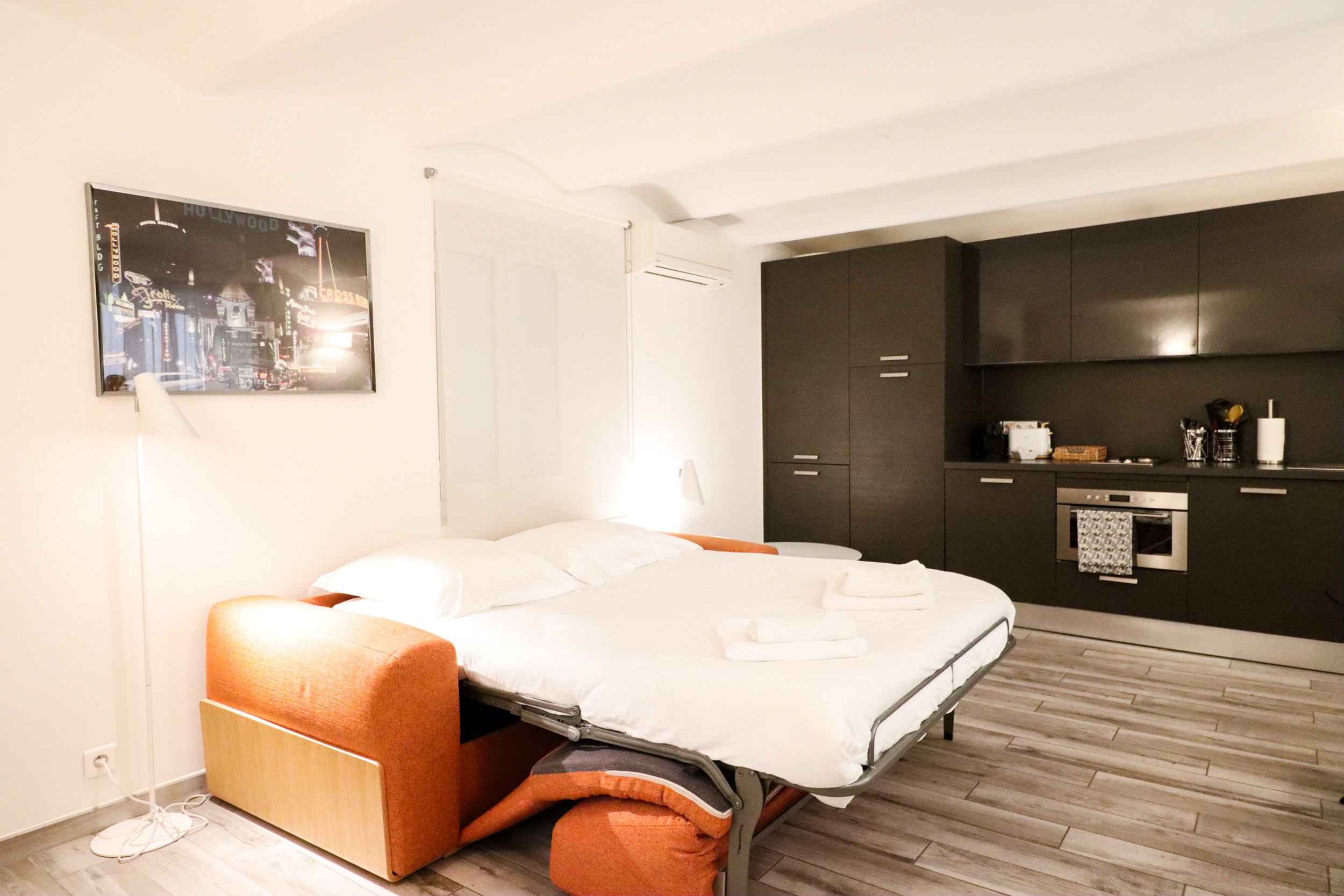 Chambre moderne avec un lit fait, deux serviettes pliées, un fauteuil orange, un meuble de rangement noir, une fenêtre, et un cadre avec une image de la ville la nuit.