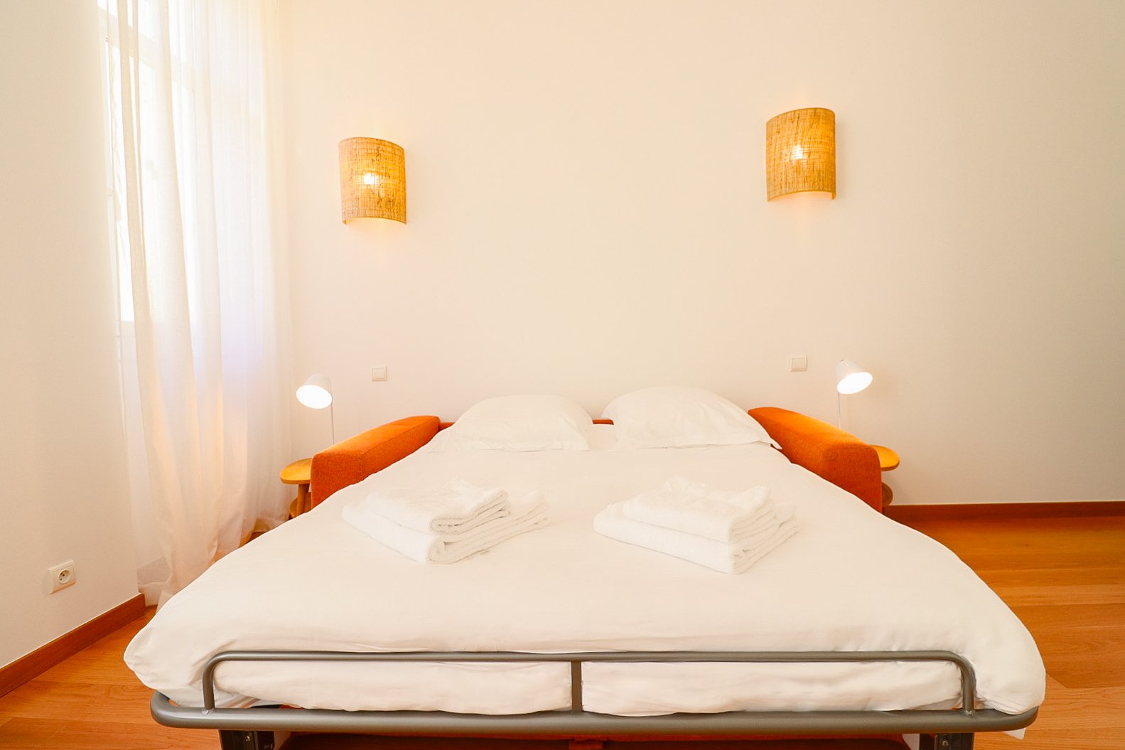 Chambre simple avec un grand lit double, deux lampes de lecture à chaque côté, deux lampes murales, linge de lit blanc, deux serviettes pliées sur le lit, rideaux transparents devant une fenêtre.