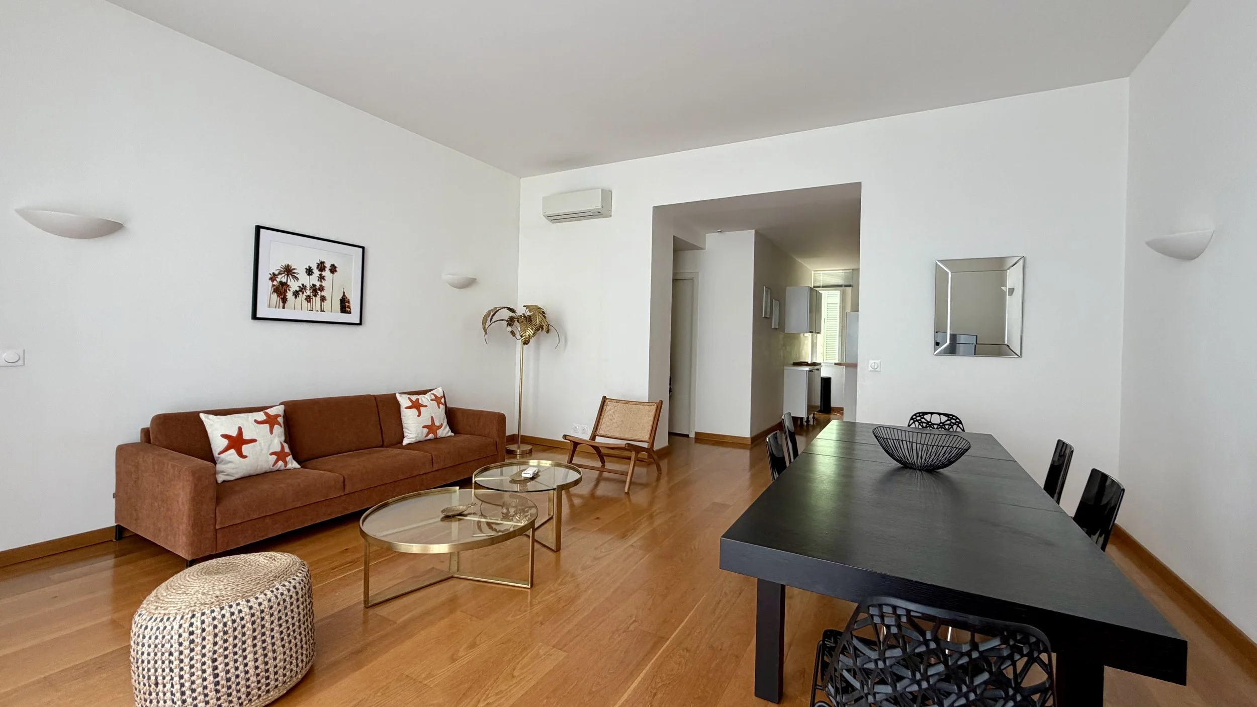 Salon intérieur avec un canapé marron, des coussins étoile de mer, une table basse en verre, un fauteuil en bois, un lampadaire doré, une plante verte, un tableau avec des palmiers, et une table à manger noire avec des chaises