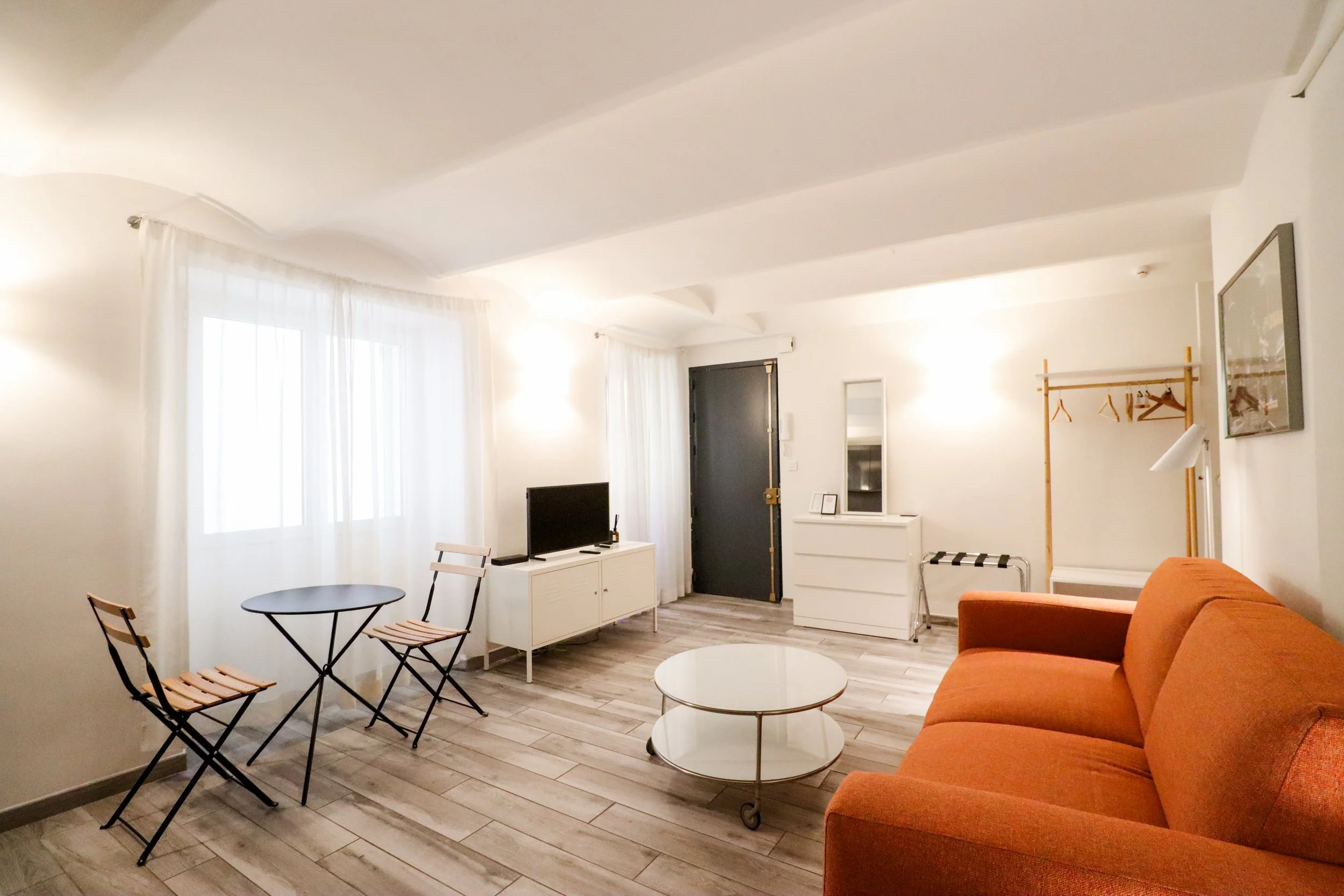 Intérieur d'un salon avec canapé orange, table basse ronde, télévision, fauteuils en bois pliants, grande fenêtre avec rideaux blancs, murs blancs, décor minimaliste.