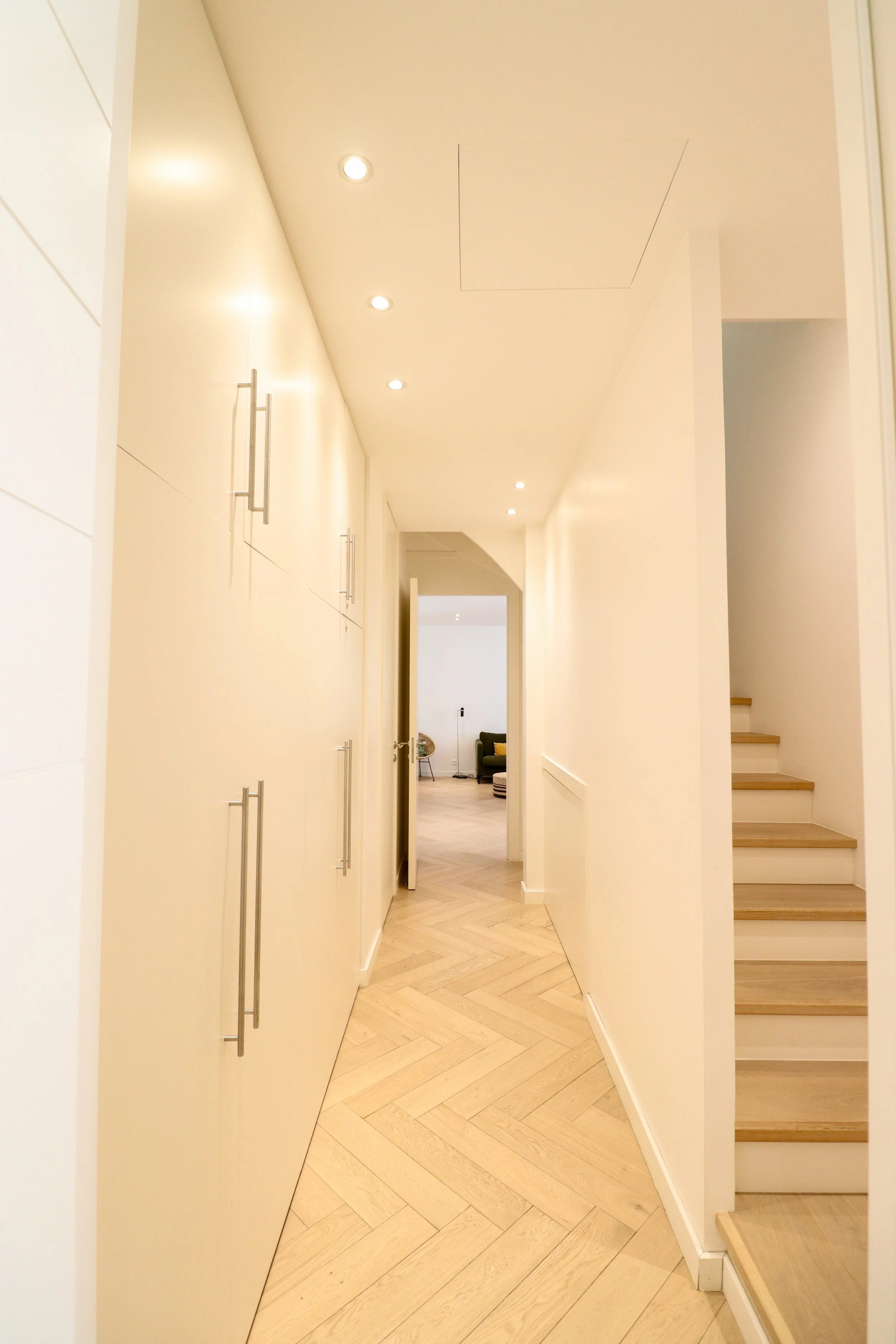 le couloir d'une maison ou d'un appartement avec un parquet clairt et des murs blancs, un escalier en bois, et des armoires de rangement blanches avec des poignées métalliques.
