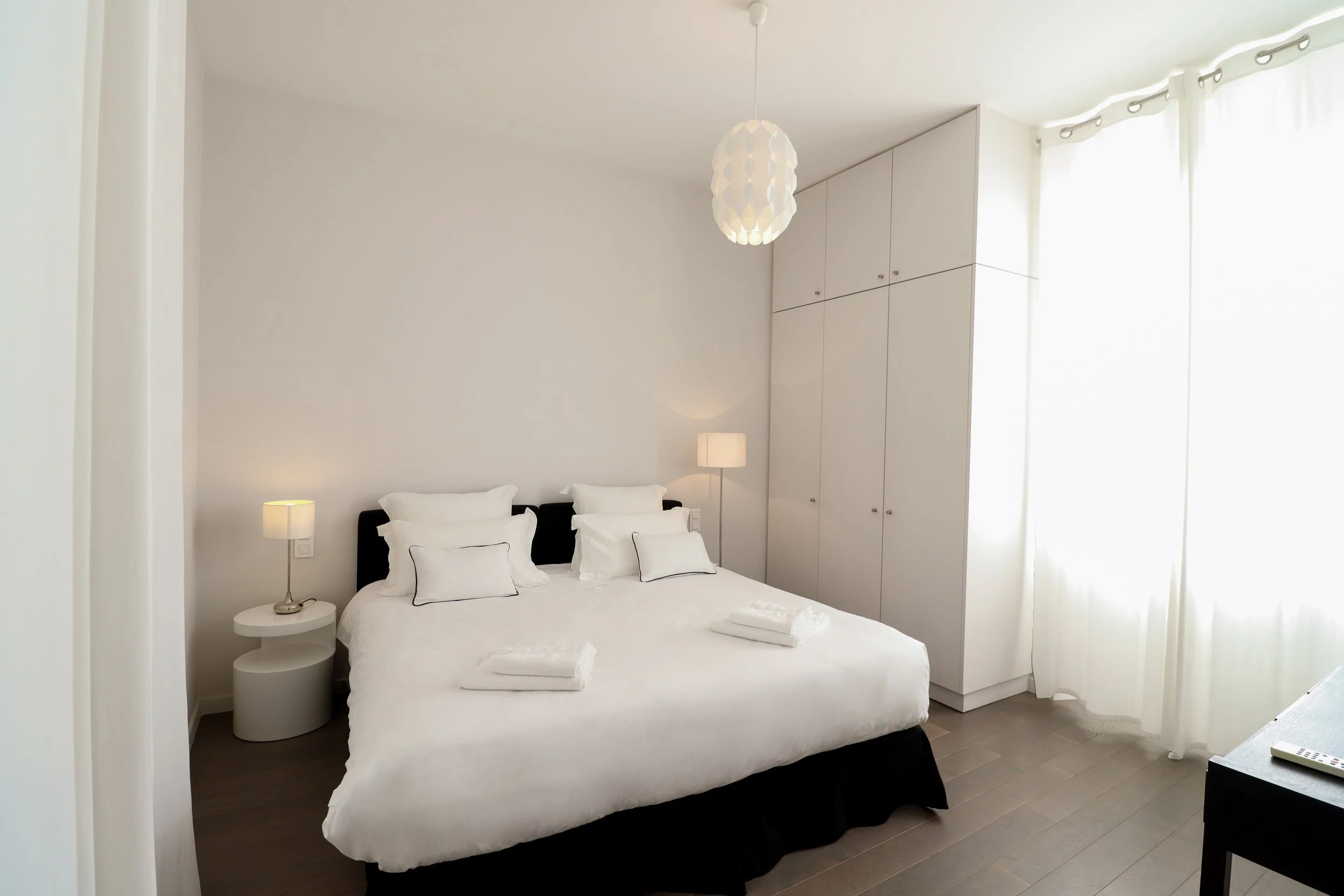 Chambre à coucher avec un lit blanc, deux lampes de chevet, des rideaux blancs et des armoires blanches intégrées.