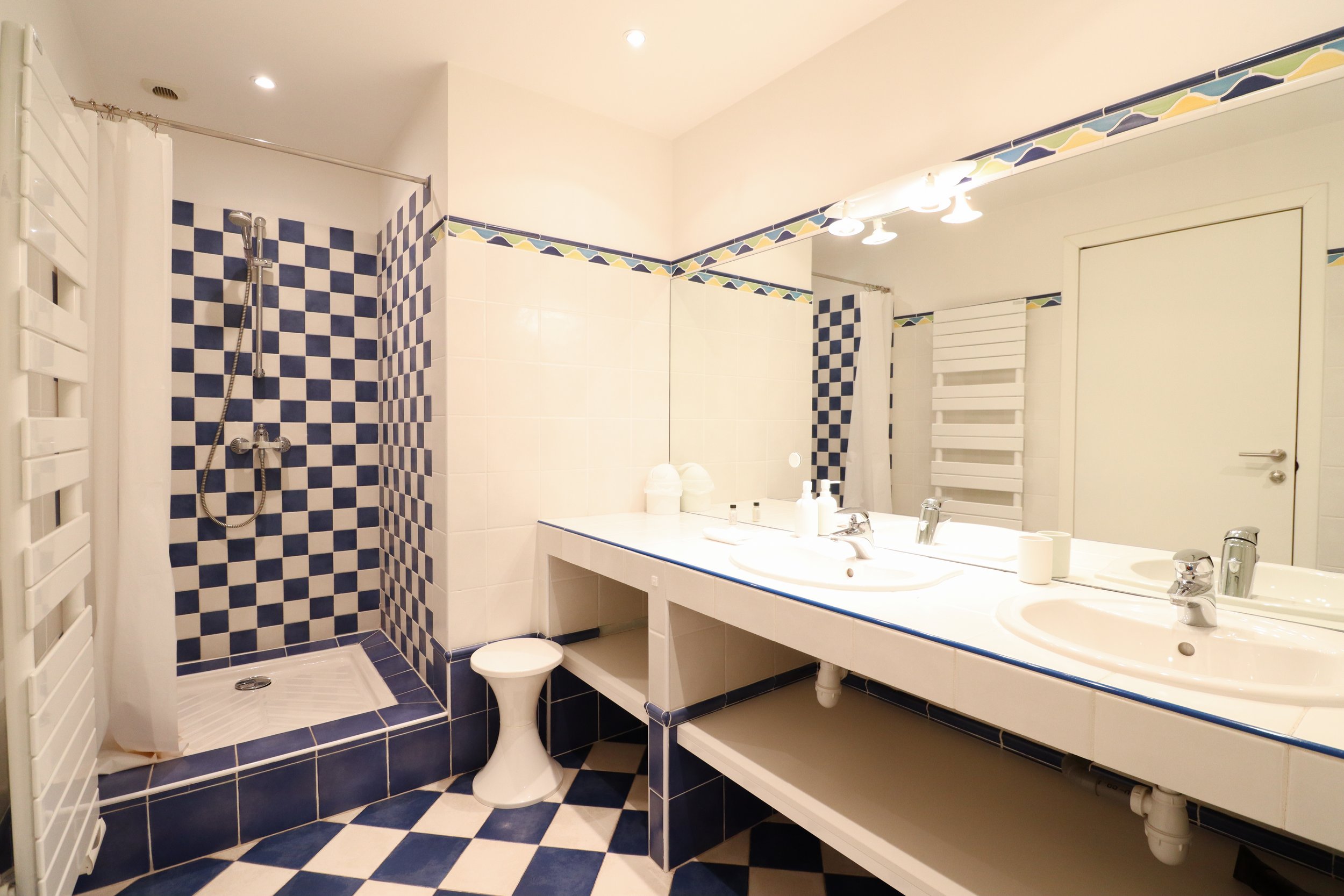 Vue de salle de bain avec douche, double lavabo, grands miroirs, et décoration en carrelage bleu et blanc.