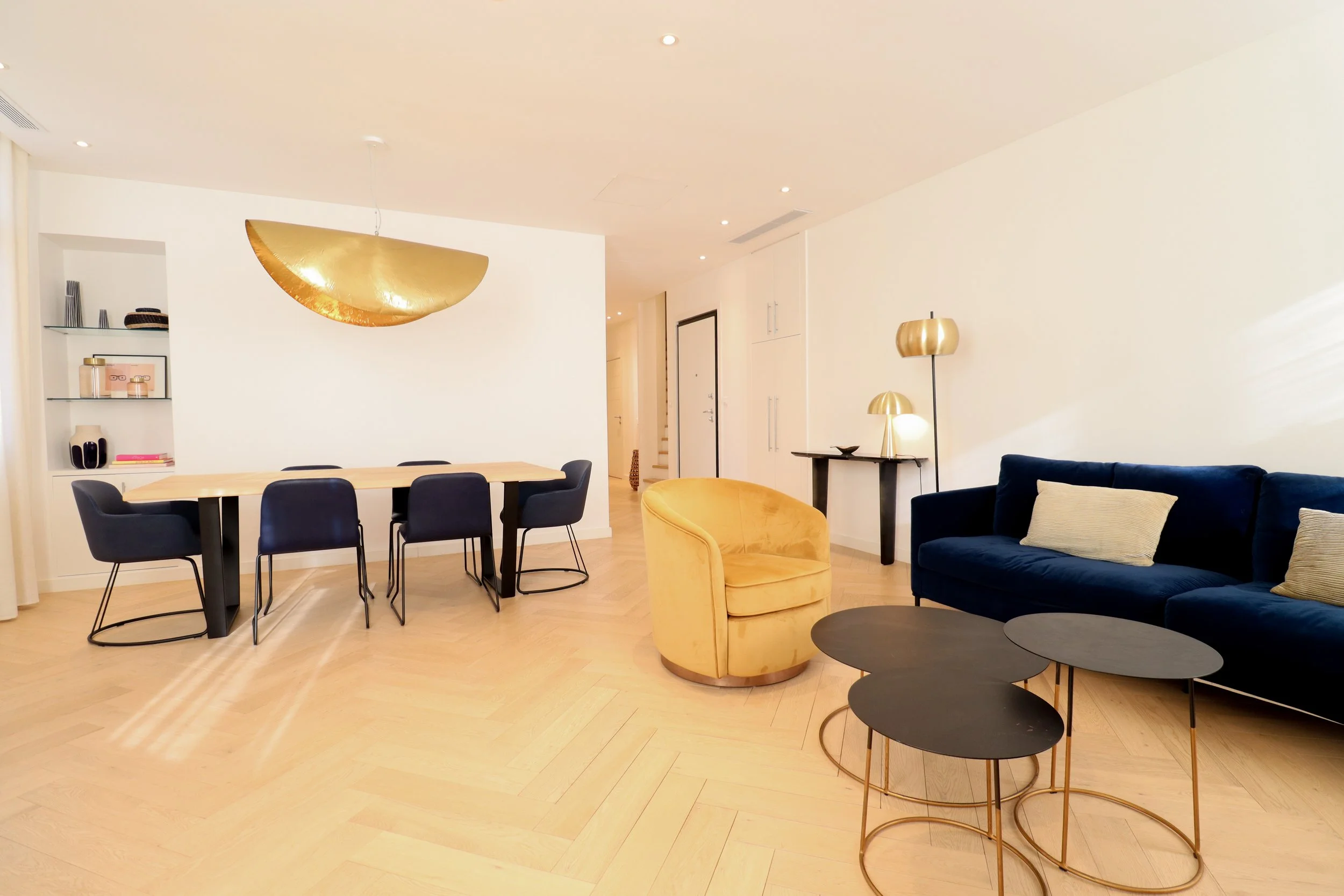 Salon moderne avec canapé bleu, fauteuil jaune, table d'appoint noire, lampe dorée, décoration élégante, murs blancs et sol en bois clair.
