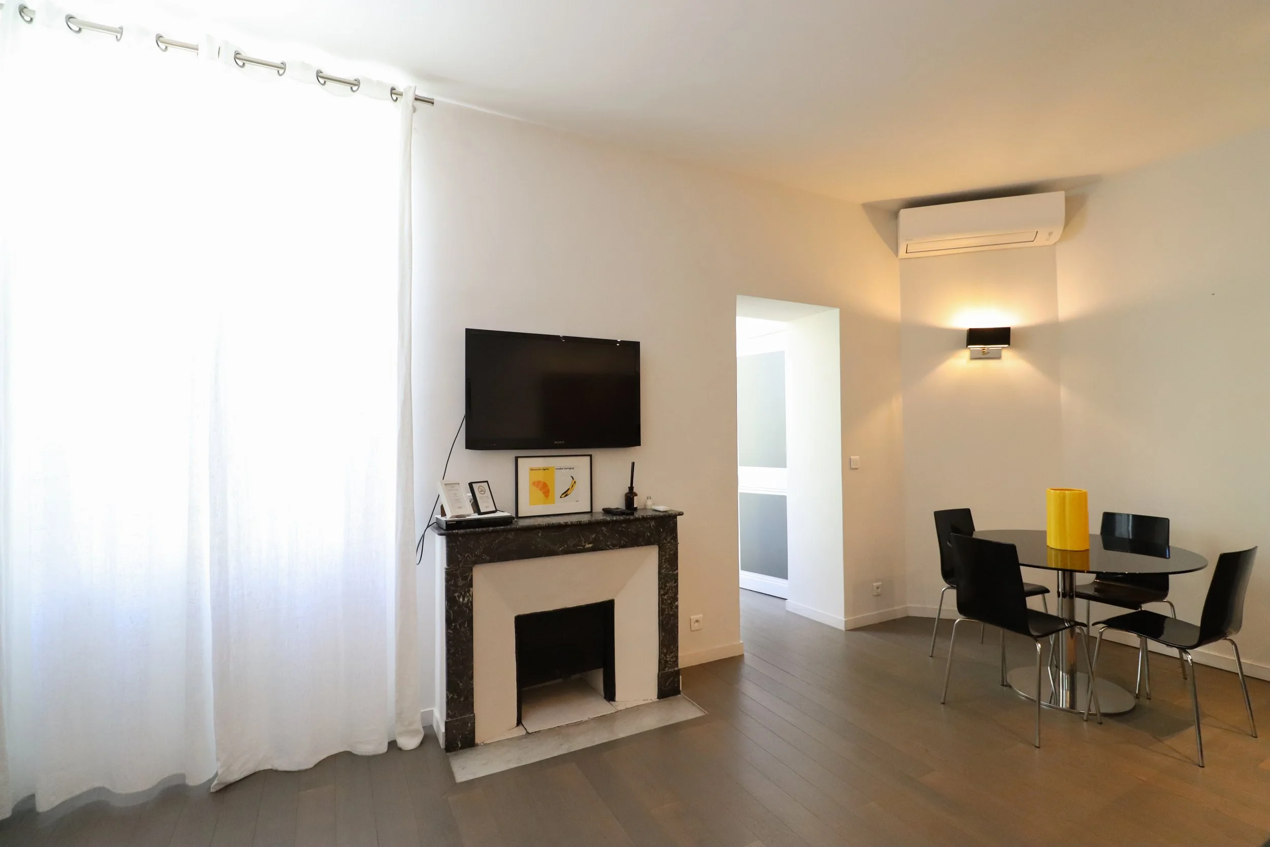 Intérieur d'un salon moderne avec un mur blanc, une télévision, une cheminée en marbre, un tableau encadré, des meubles noirs, une table ronde noire avec des chaises noires, et un vase jaune sur la table, éclairé par un lampadaire et une lumière mura
