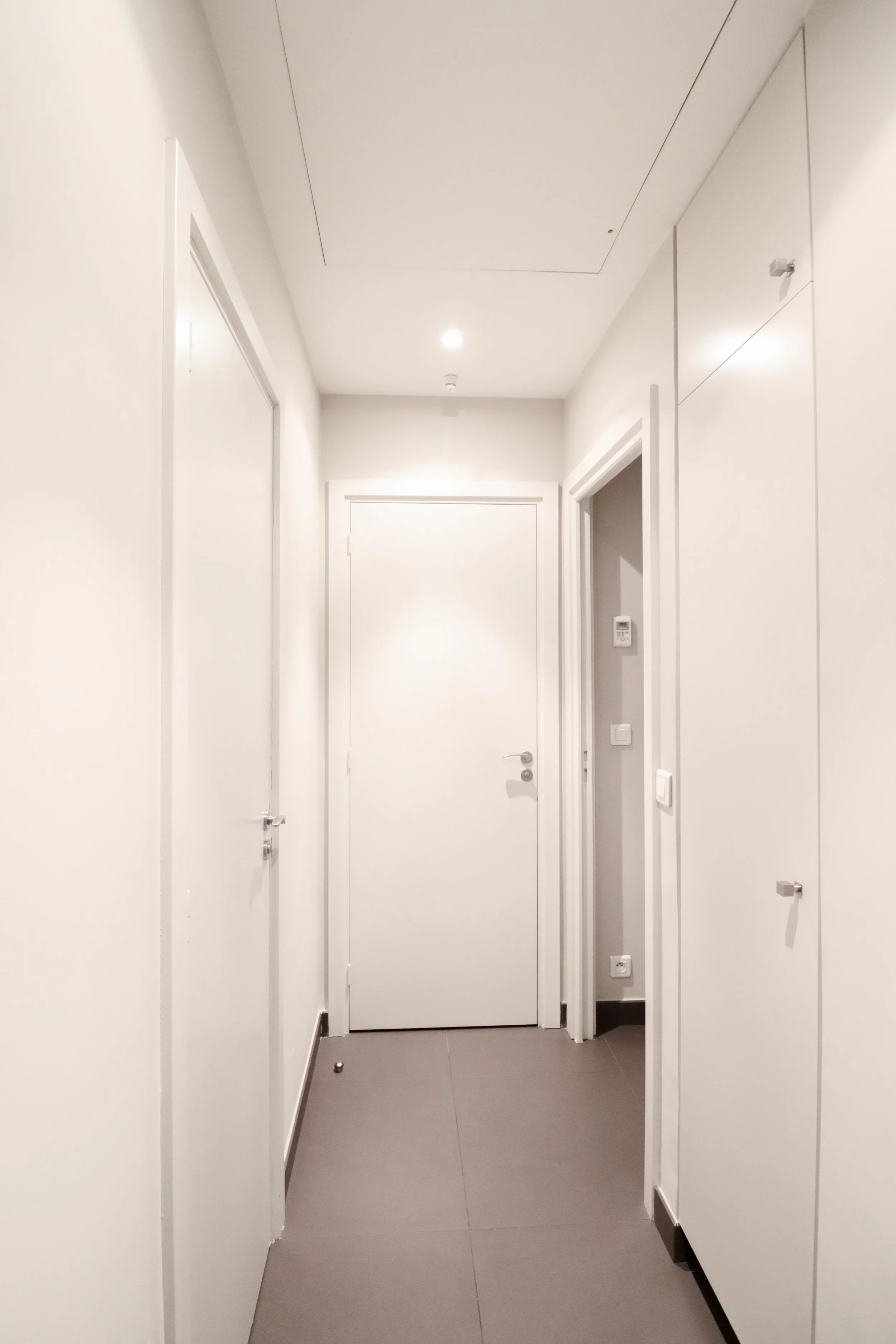 Couloir blanc avec portes fermées, plafond lumineux et sol en carrelage gris, et un panneau de contrôle à droite.