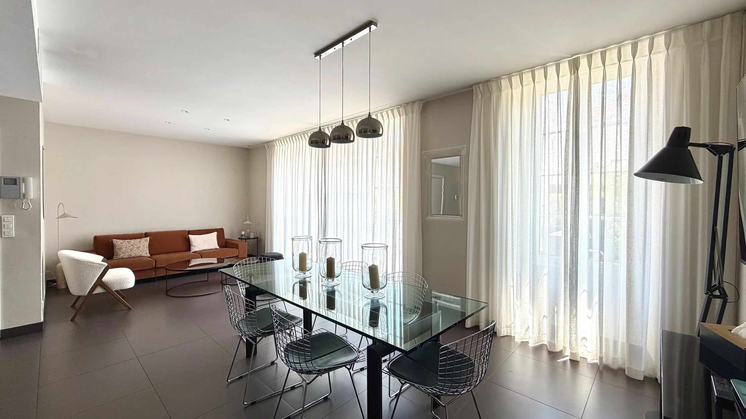 Salon moderne avec grande baie vitrée, rideaux blancs, canapé marron, fauteuil blanc, table en verre avec bougies, lampadaire noir, suspension noire