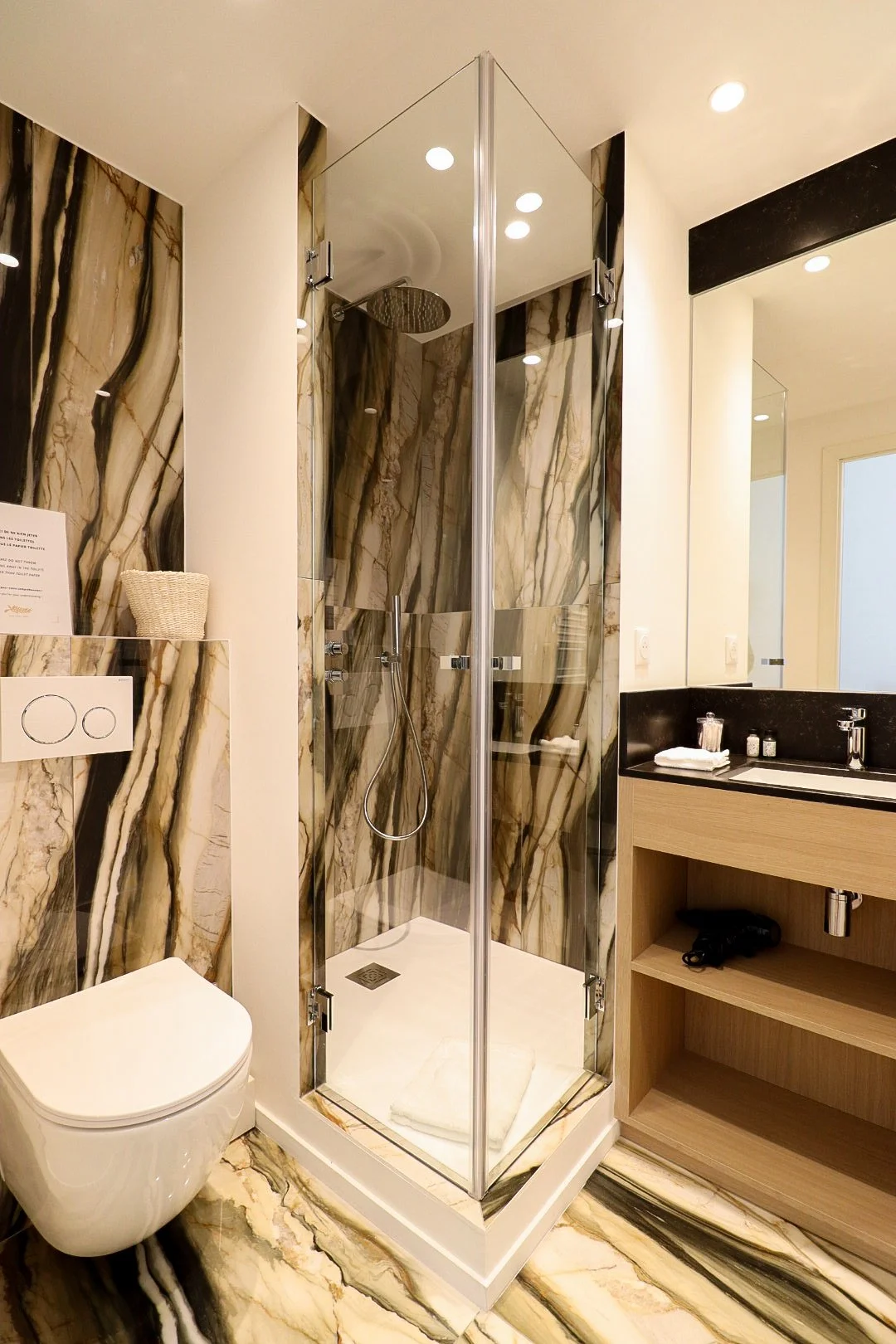 Salle de bain moderne avec mur en marbre, cabine de douche en verre, toilette suspendu et lavabo avec miroir et rangement en bois.