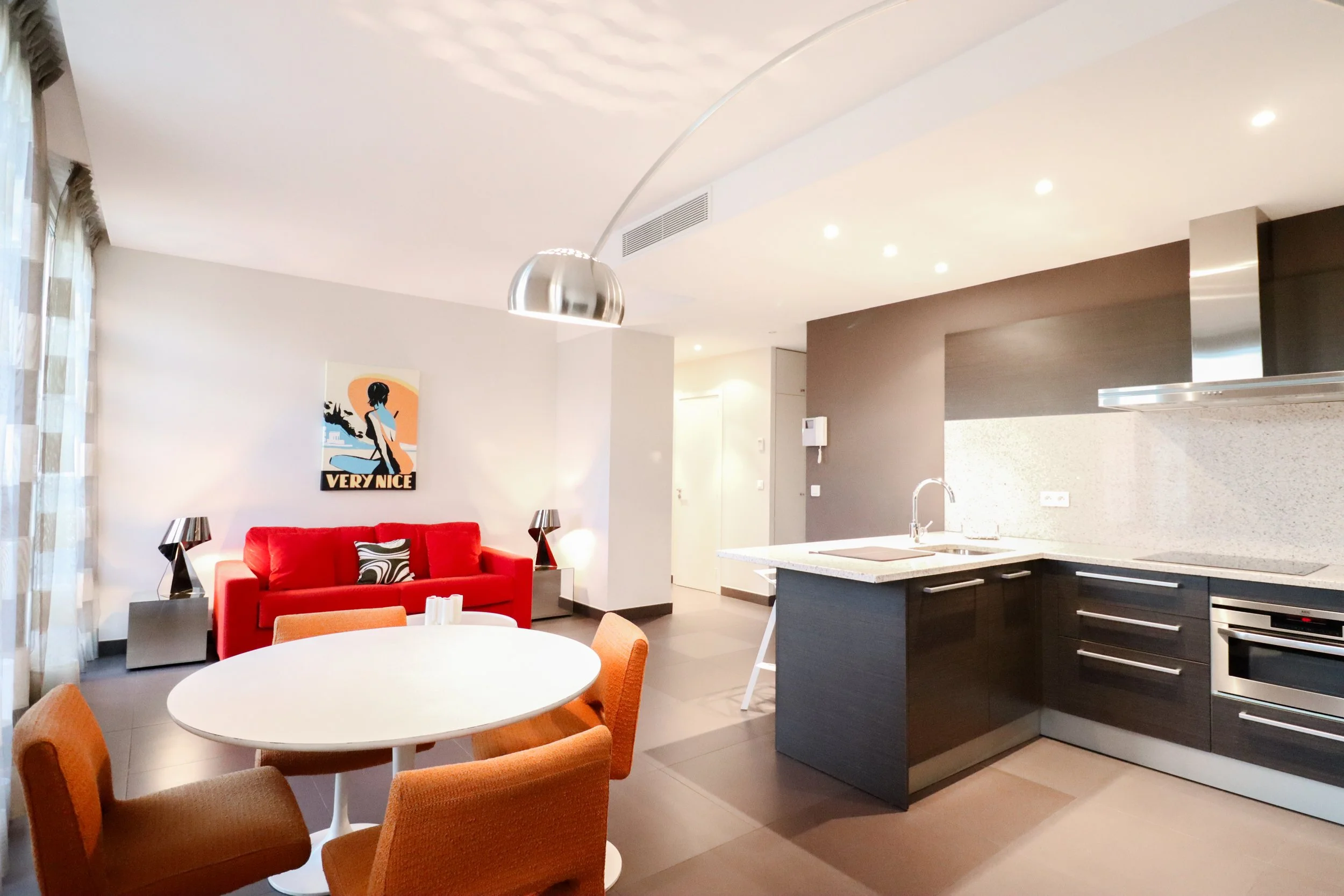 Intérieur moderne comprenant un salon avec un canapé rouge, un tableau mural avec la phrase "Very Nice", un mobilier en acier et une cuisine en arrière-plan avec un plan de travail en quartz clair, des appareils électroménagers en acier inoxydable, et un éclairage encastré au plafond.